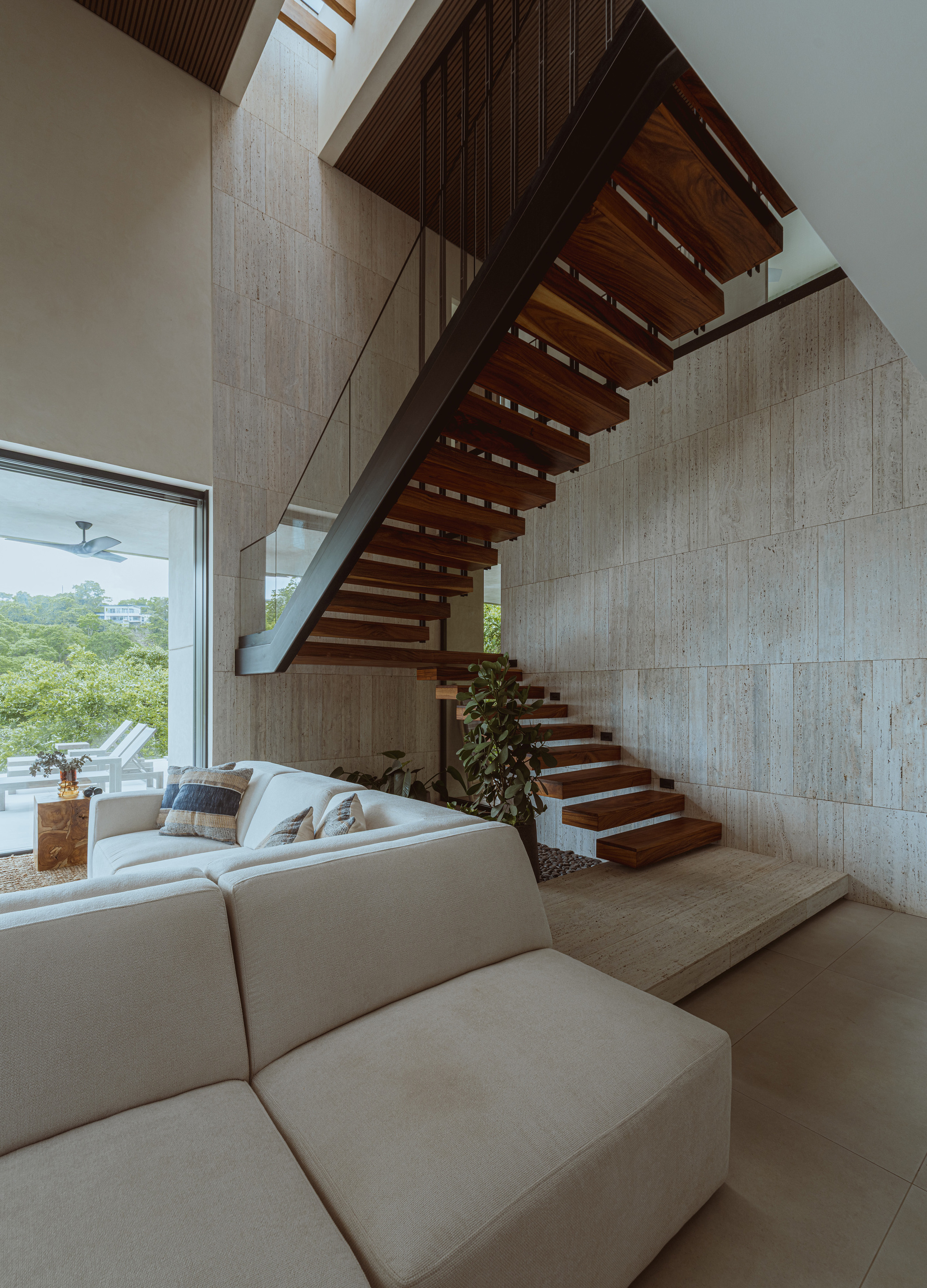 Floating timber-tread staircase against a travertine wall, with a pale sectional sofa in front and green hillside through the window