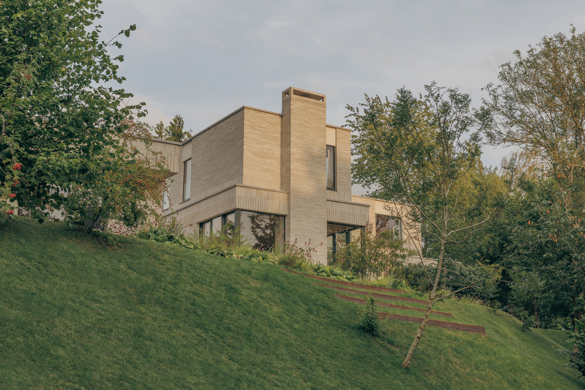 Pale brick house with a tall chimney stack set above a grassy slope with weathered steel steps at dusk