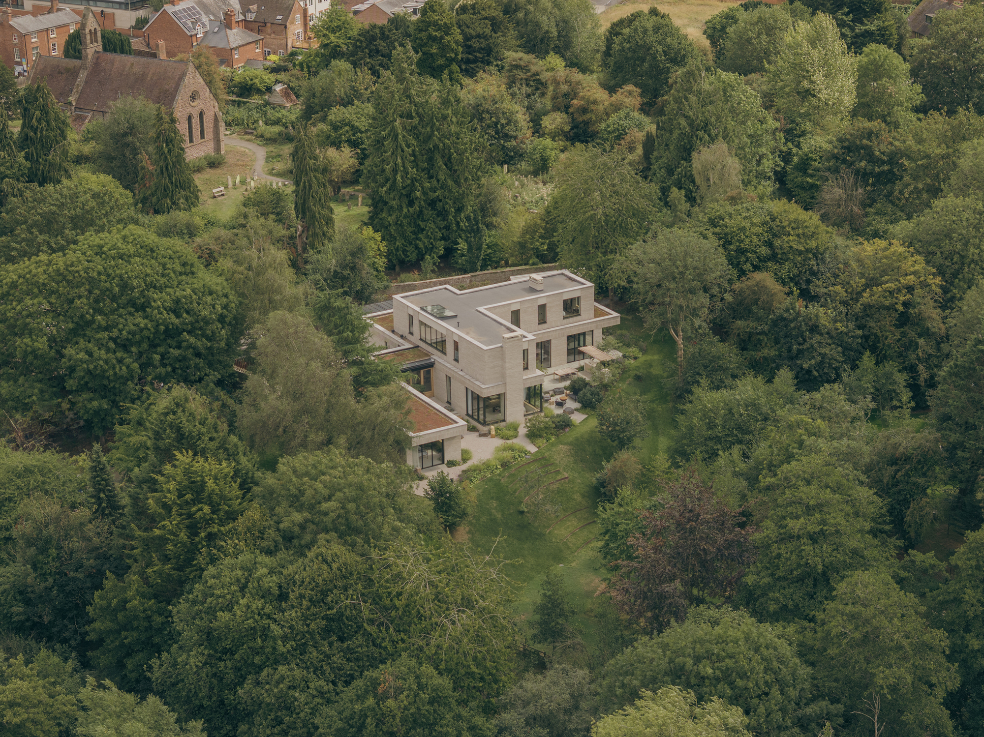 Aerial view of the pale brick house with flat roof and terrace, surrounded by dense woodland and a church beyond