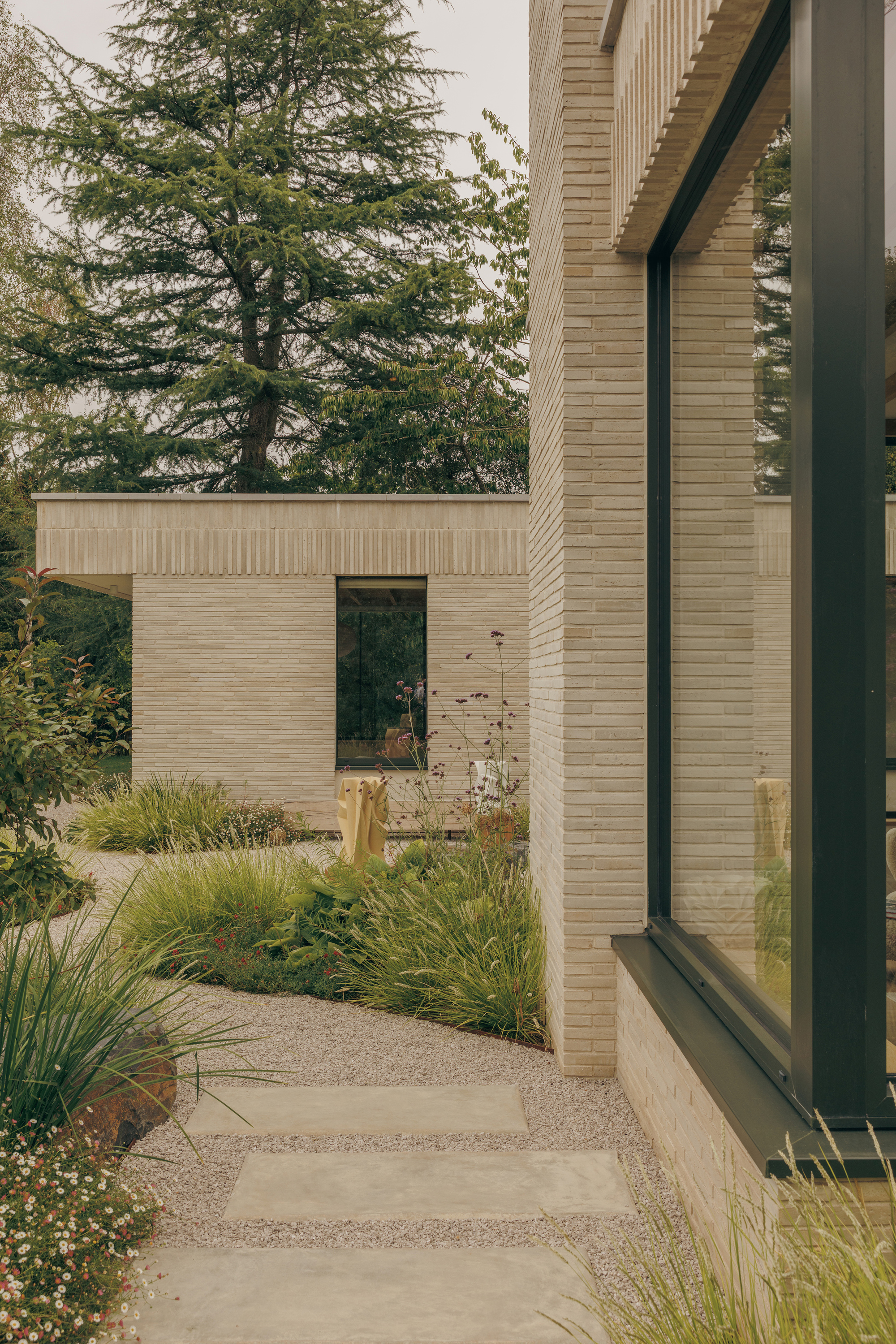 Slim pale brick walls and dark framed glazing beside a gravel path with stepping stones and grasses under a cedar