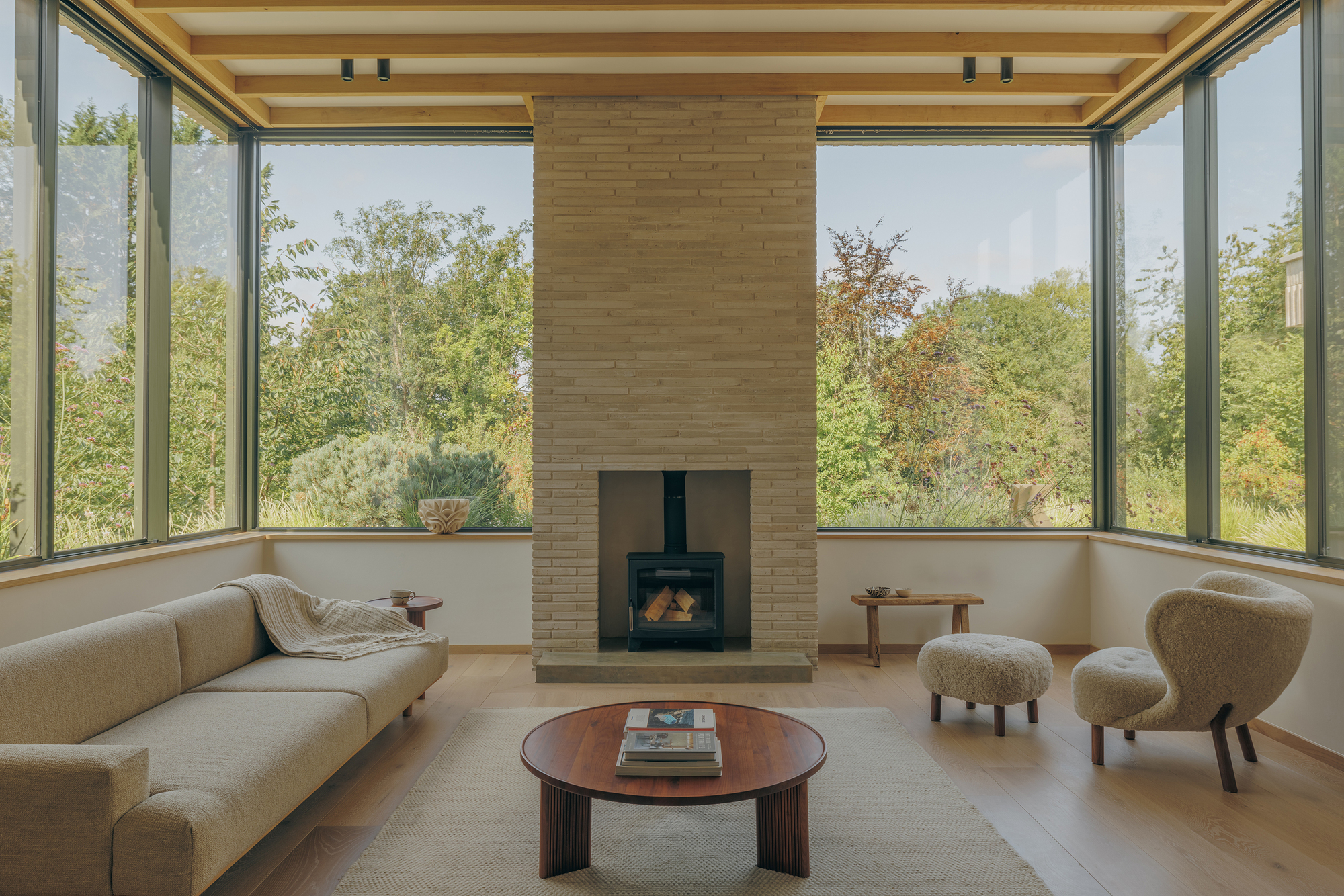 Living room with brick chimney and wood stove between corner windows, framed by trees in soft daylight