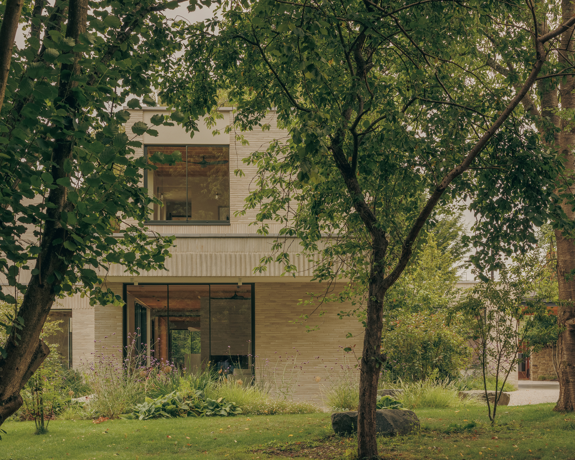 Two-storey brick facade with large windows seen through garden trees and a boulder on the lawn in summer
