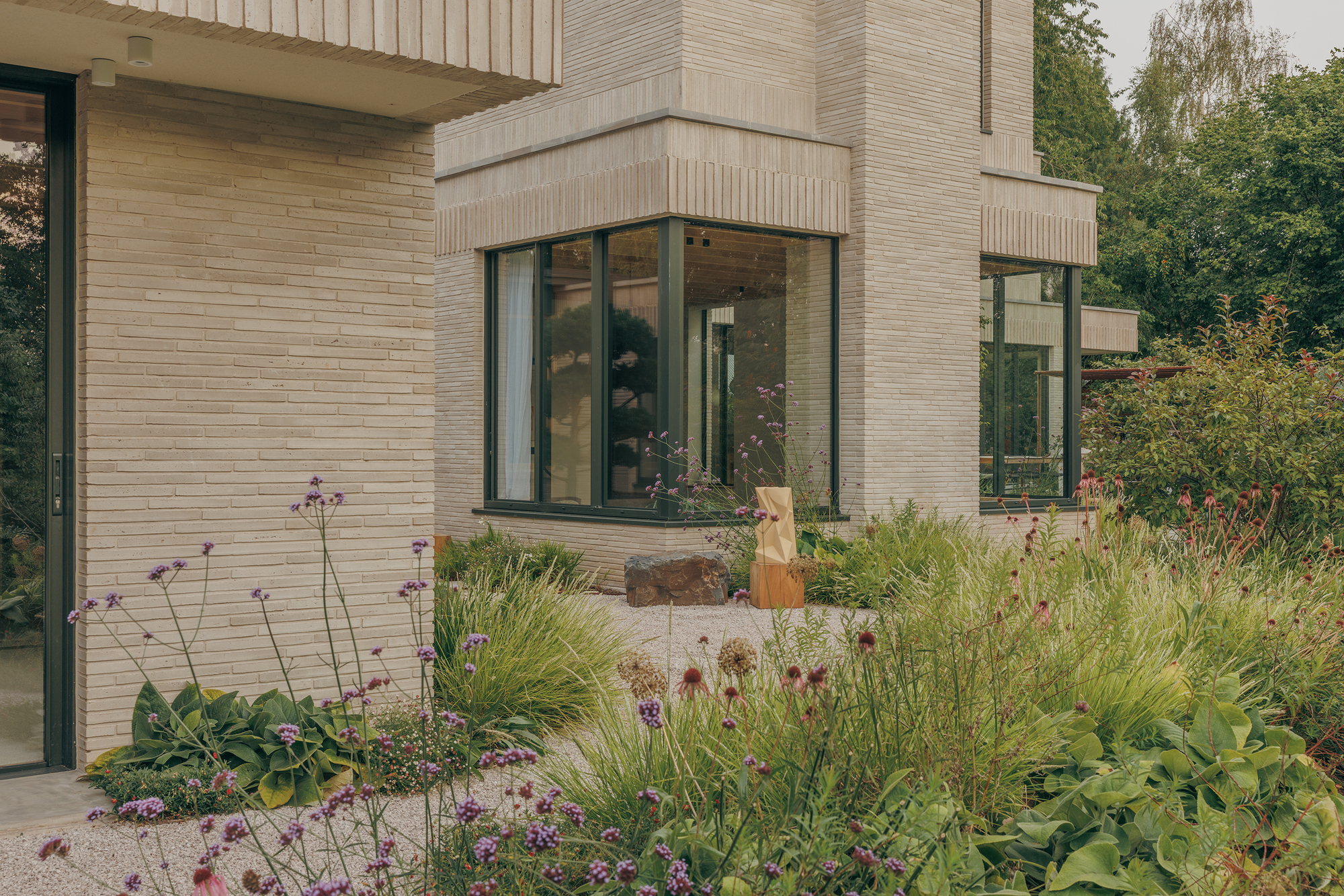 Pale brick corner with a dark-framed bay window above a gravel garden of purple verbena and grasses in overcast light