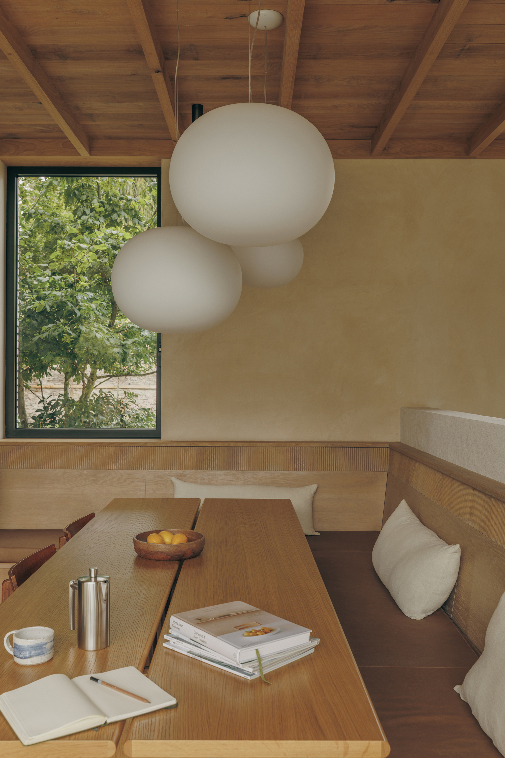 Dining nook with built-in oak banquette, clustered white globe pendants and a window onto green foliage