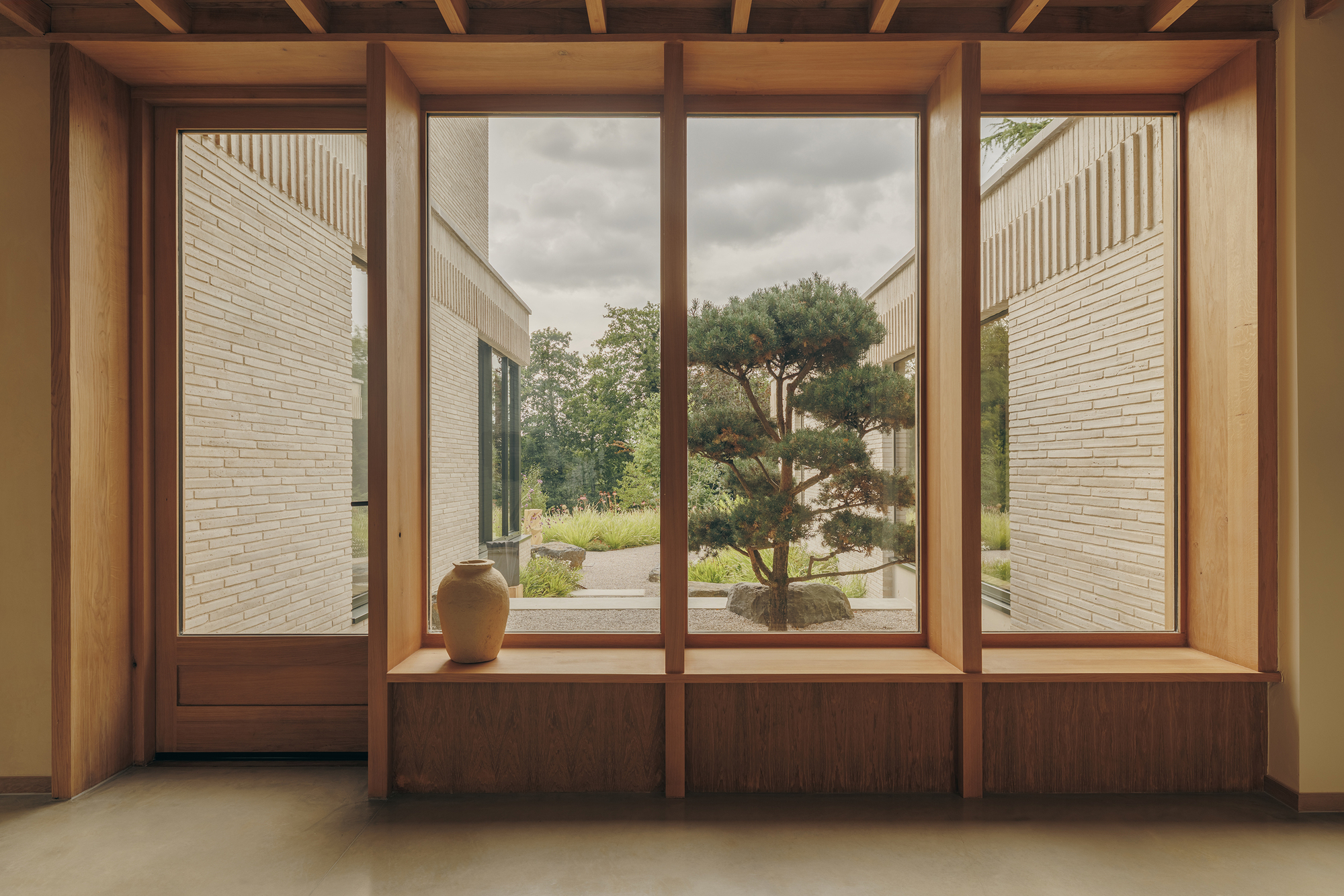 Timber-framed window seat with a ceramic vase, looking onto a sculpted pine and brick wings under cloudy sky