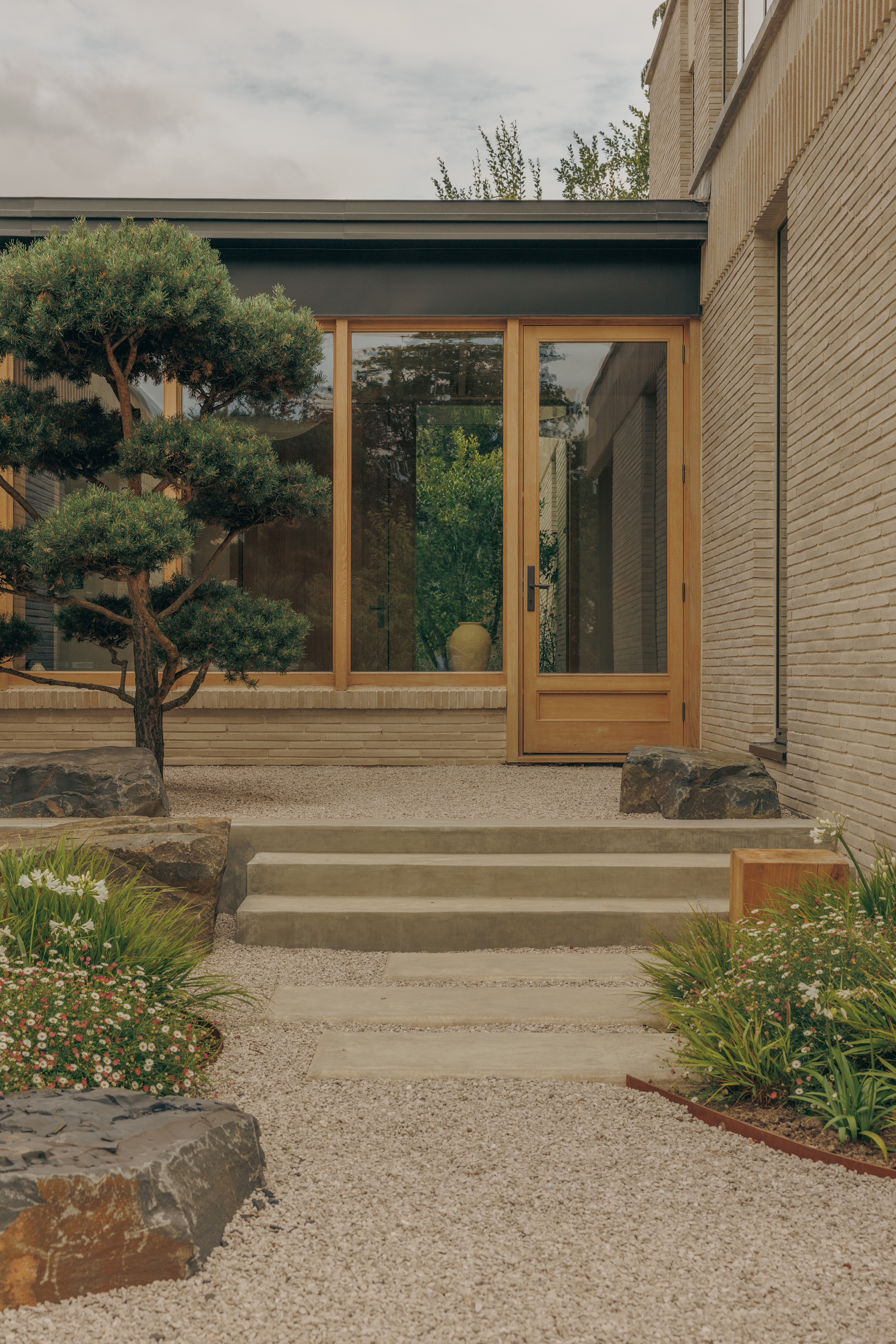 Concrete steps rising from a gravel court to a timber-framed glass door, with a clipped pine and boulders