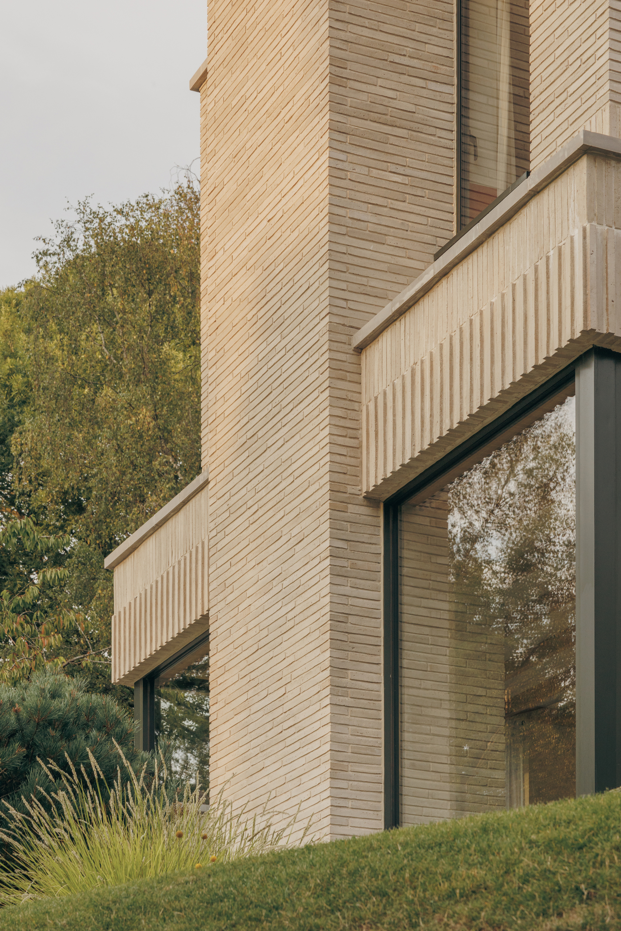 Corner of a pale slim-brick tower with ribbed concrete canopies and tall glazing, reflecting trees above a grassy bank