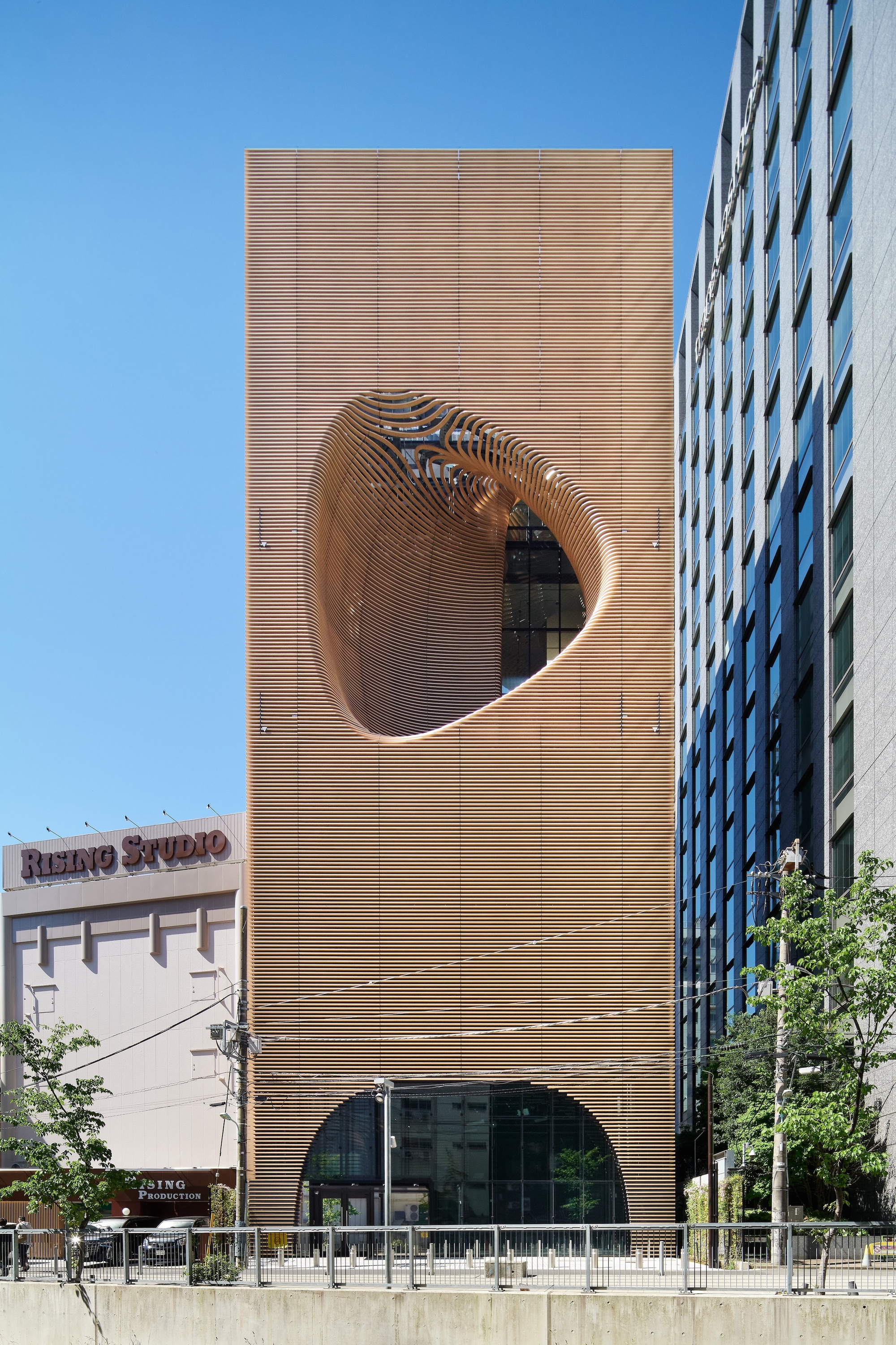 Front facade of horizontal timber louvers with a large oval cutout and arched ground-floor opening under a clear blue sky