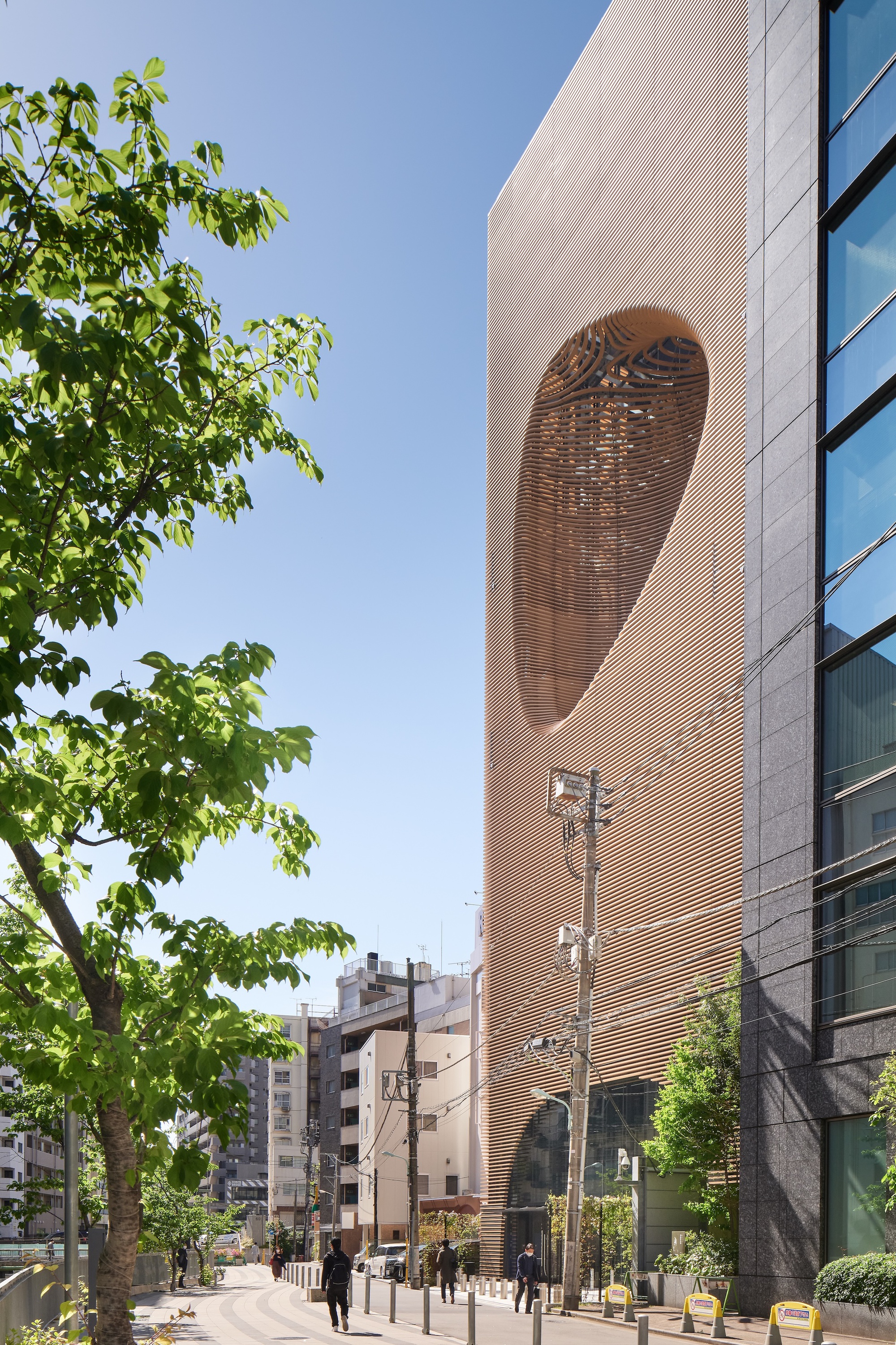 Side street view of the louvered timber facade and its oval recess, with pedestrians walking beneath a leafy tree