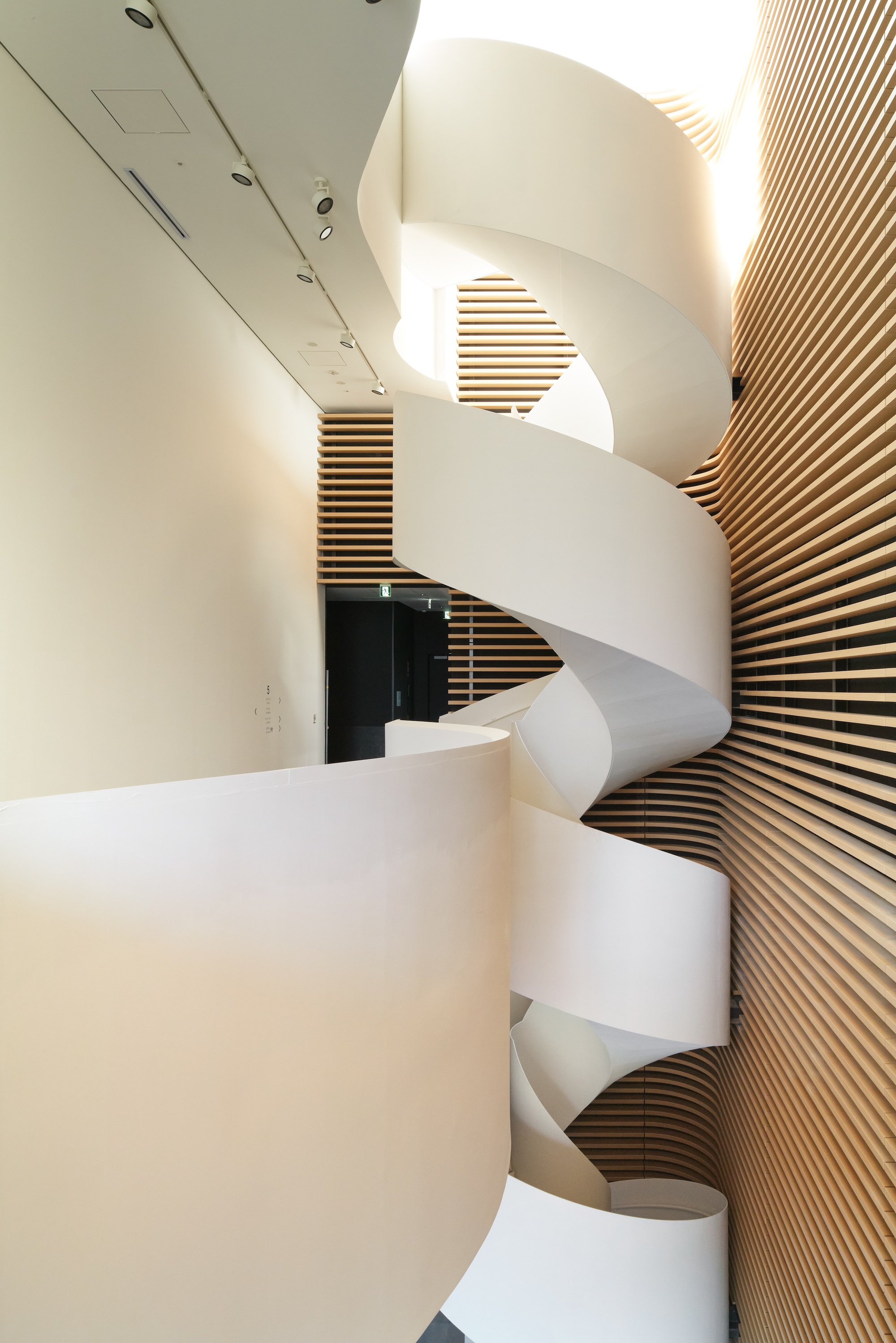 White spiral staircase winding past a timber slatted wall in a pale interior lit from above