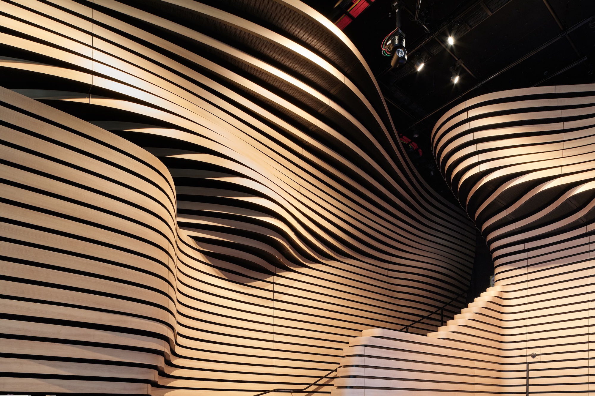 Interior wall of layered wavy timber slats rising to a dark ceiling with spotlights and a stepped ledge