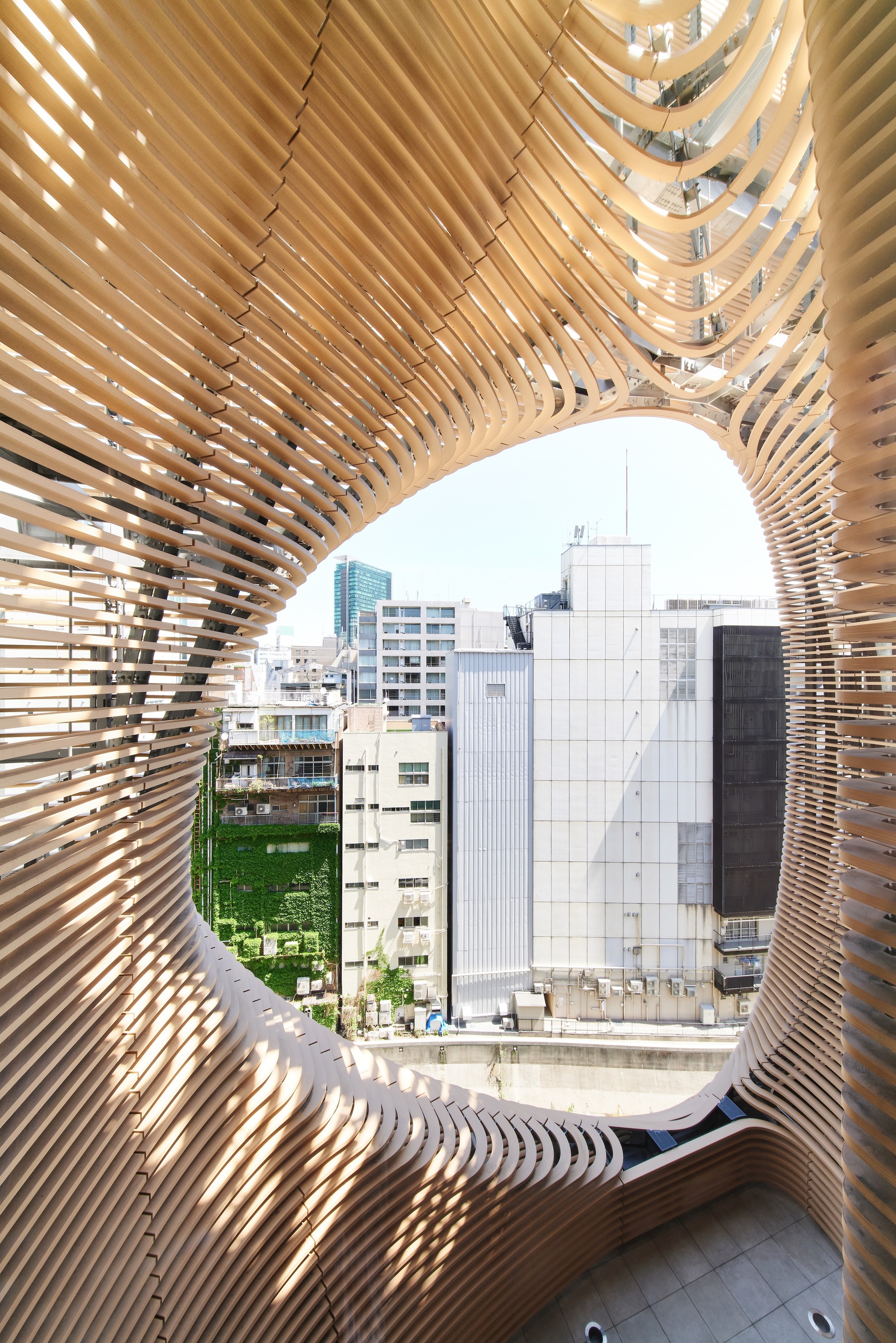 View through an oval opening in the layered timber fins toward neighboring buildings and an ivy-covered wall under a bright sky
