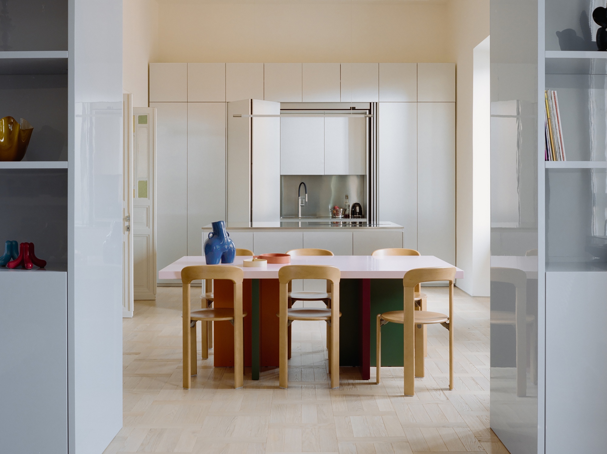Pink-topped dining table with wooden chairs and a blue vase, framed by glossy shelving, before a kitchen wall in soft daylight
