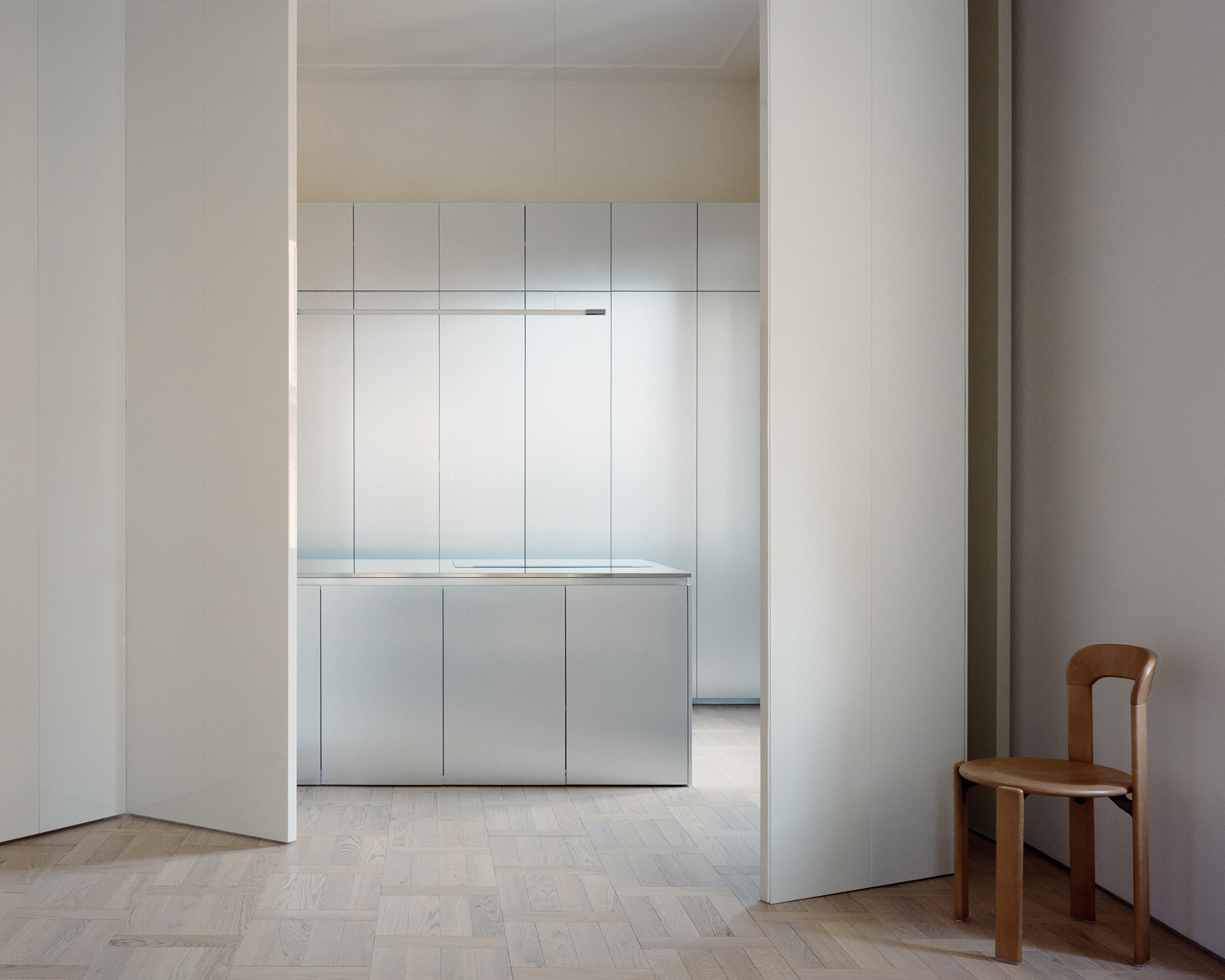 Stainless steel kitchen island and tall cabinets seen between white pivot panels, with a wooden chair beside the parquet floor