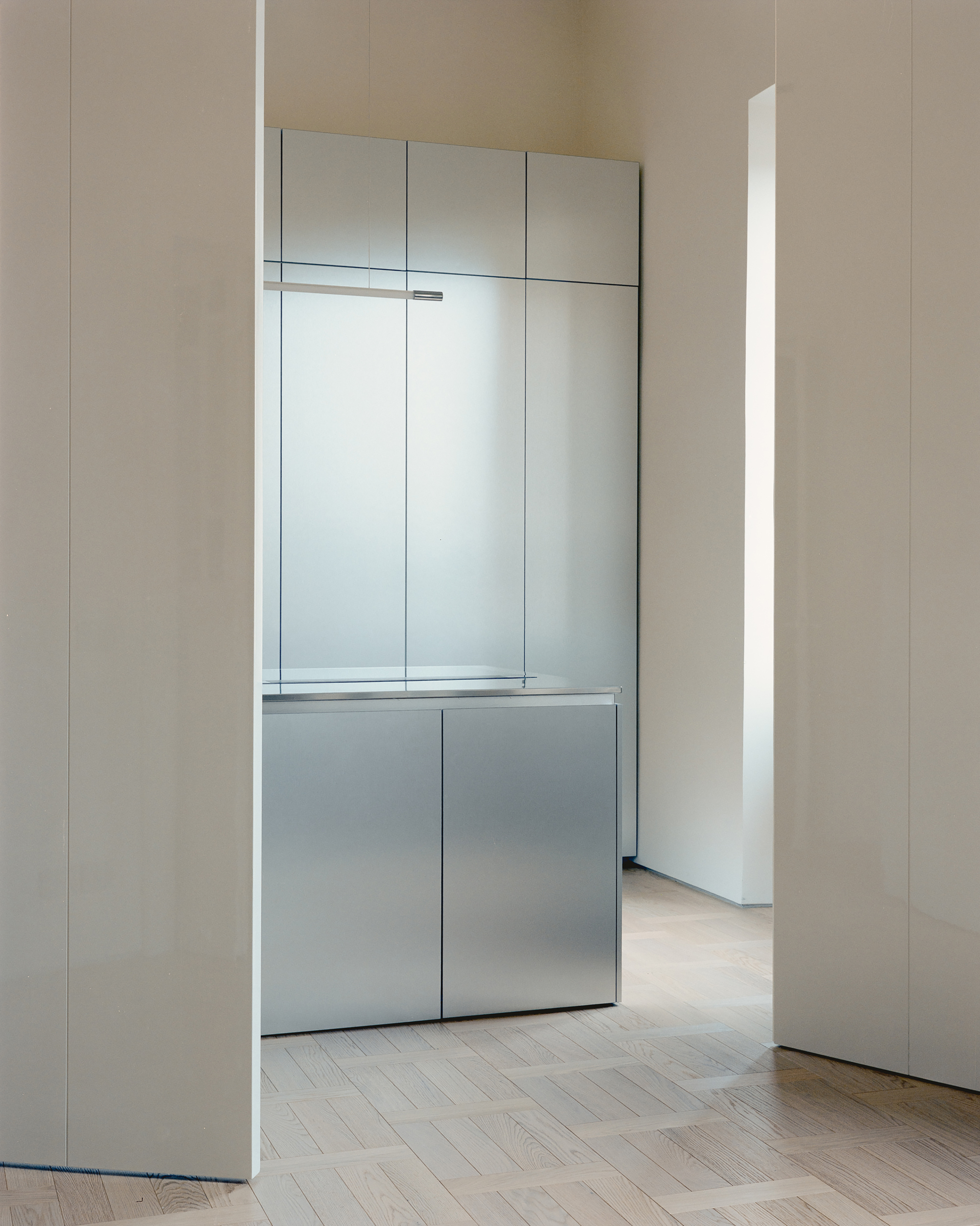 Brushed steel island and panelled cabinet wall glimpsed through a gap between tall white doors, with daylight falling on parquet