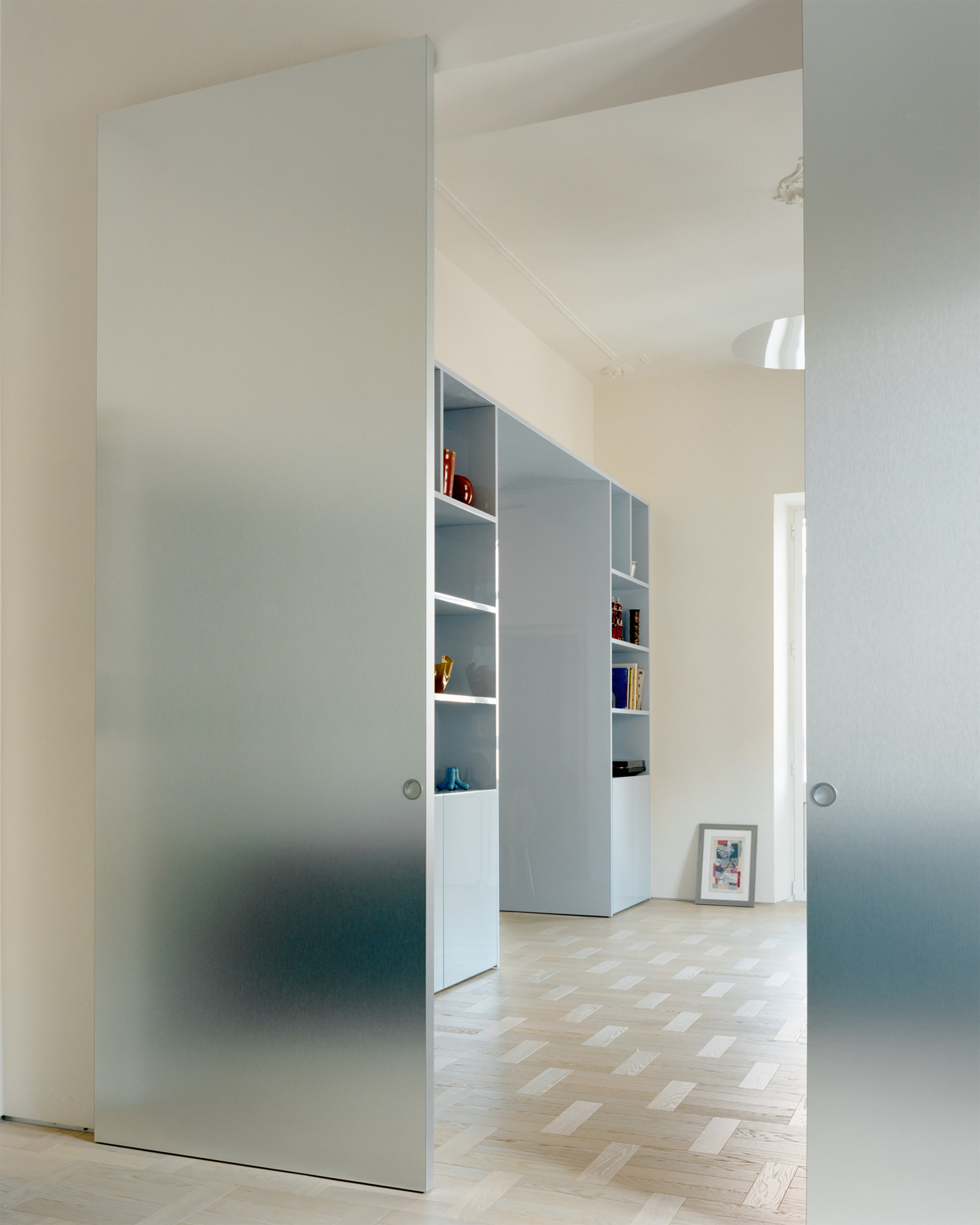 Frosted glass sliding doors open onto a pale blue shelving unit with colored objects and a framed print leaning on the floor