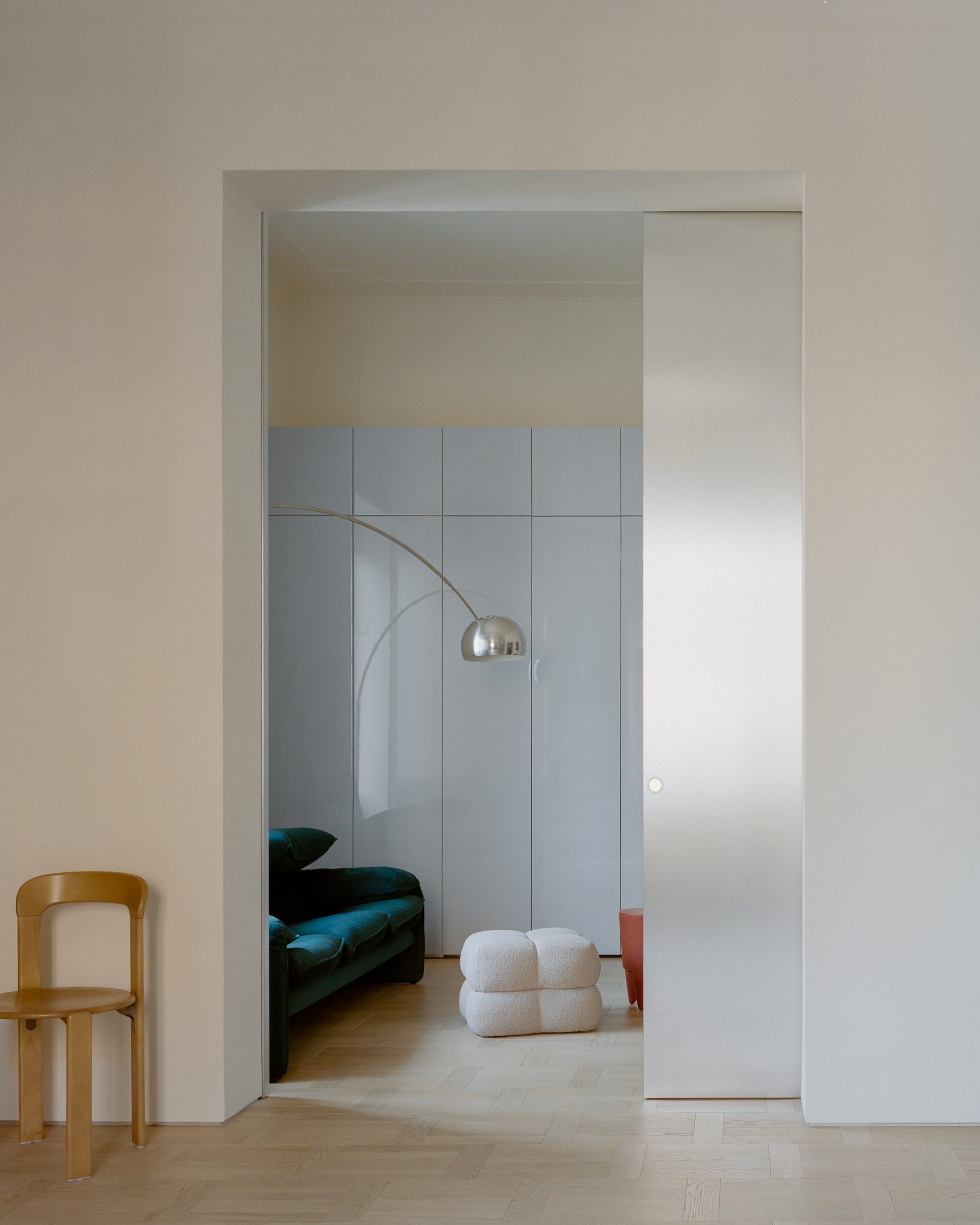 Pocket door opening onto a sitting nook with a green velvet sofa, arc floor lamp and boucle pouf, with a wooden chair at left