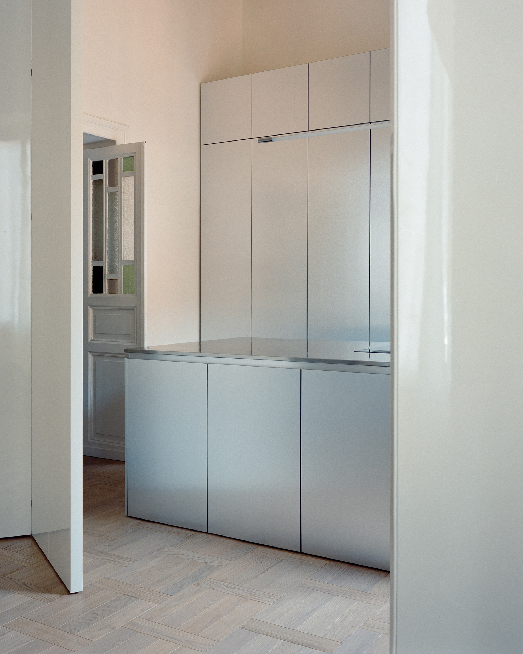 Steel island and tall cabinets beside a glazed paneled door, with soft light across herringbone parquet and a glossy white panel at right