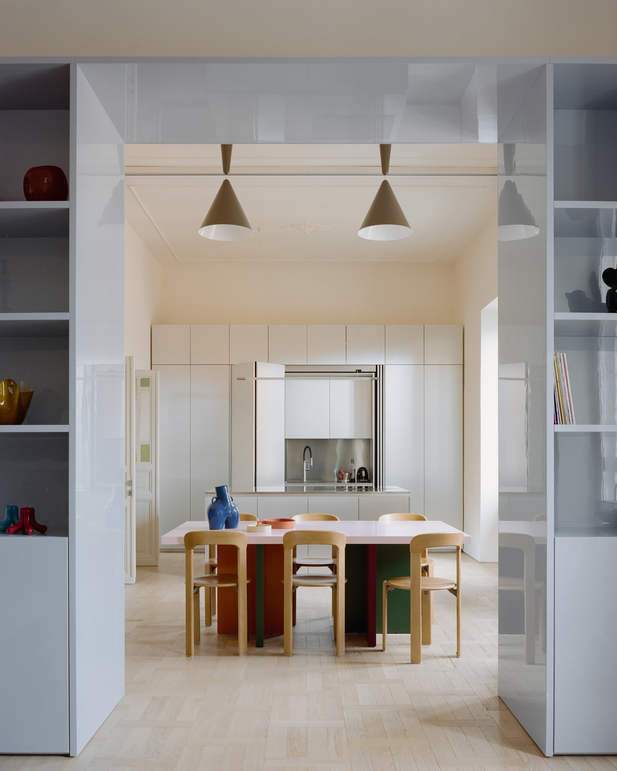Kitchen seen through a glossy pale blue shelving frame, with a pink-topped table, wooden chairs and two cone pendants in soft daylight