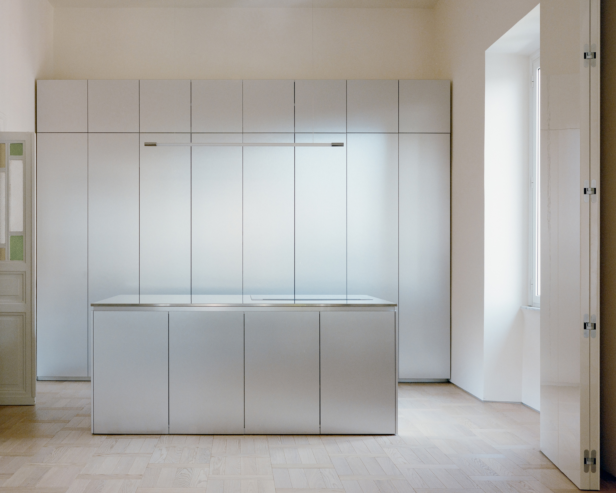 Brushed steel kitchen island facing a wall of full-height steel cabinets, with a tall window reveal lit by daylight at right