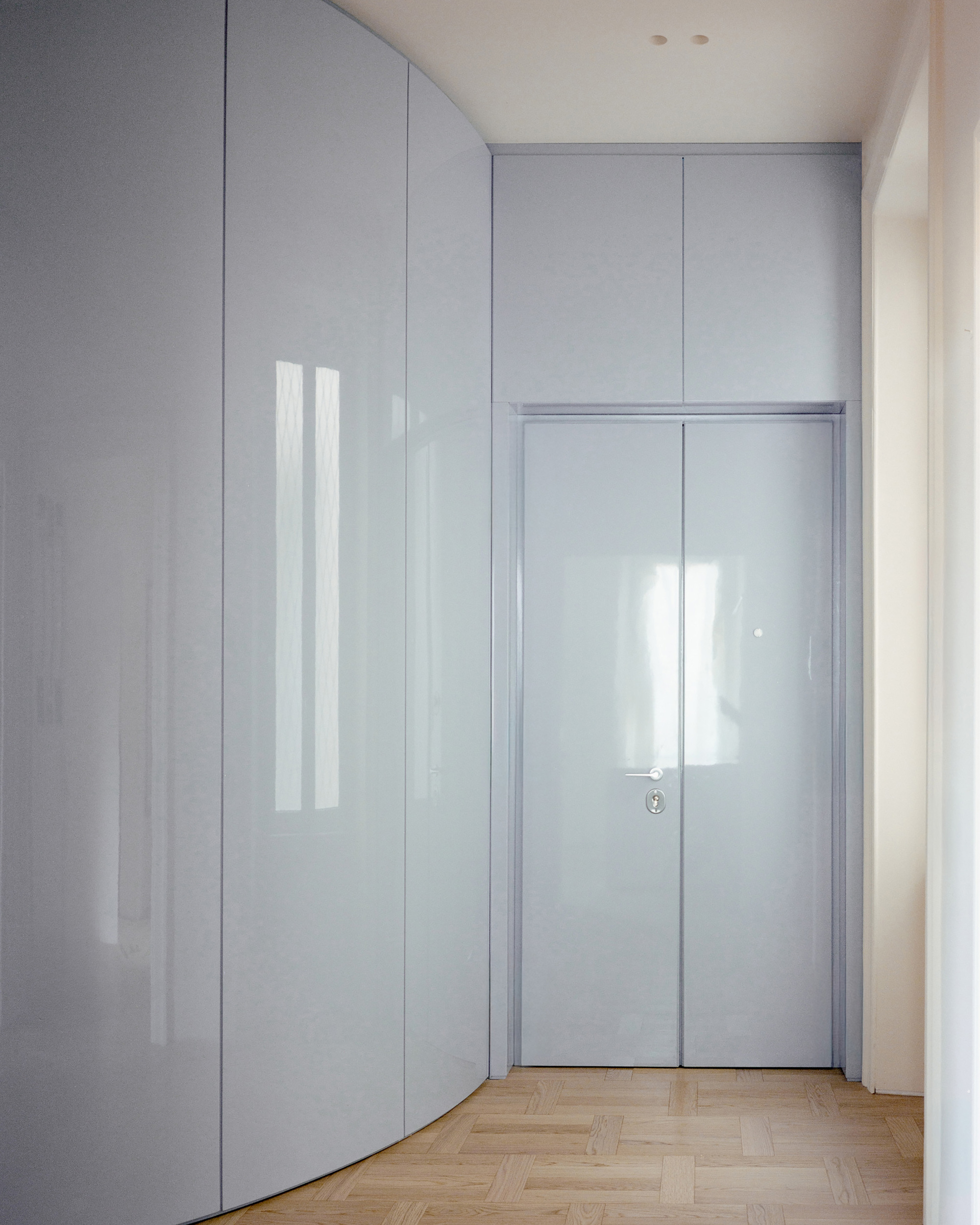 Curved glossy grey-blue cabinet wall beside a double door with a metal handle, reflecting window light on herringbone parquet
