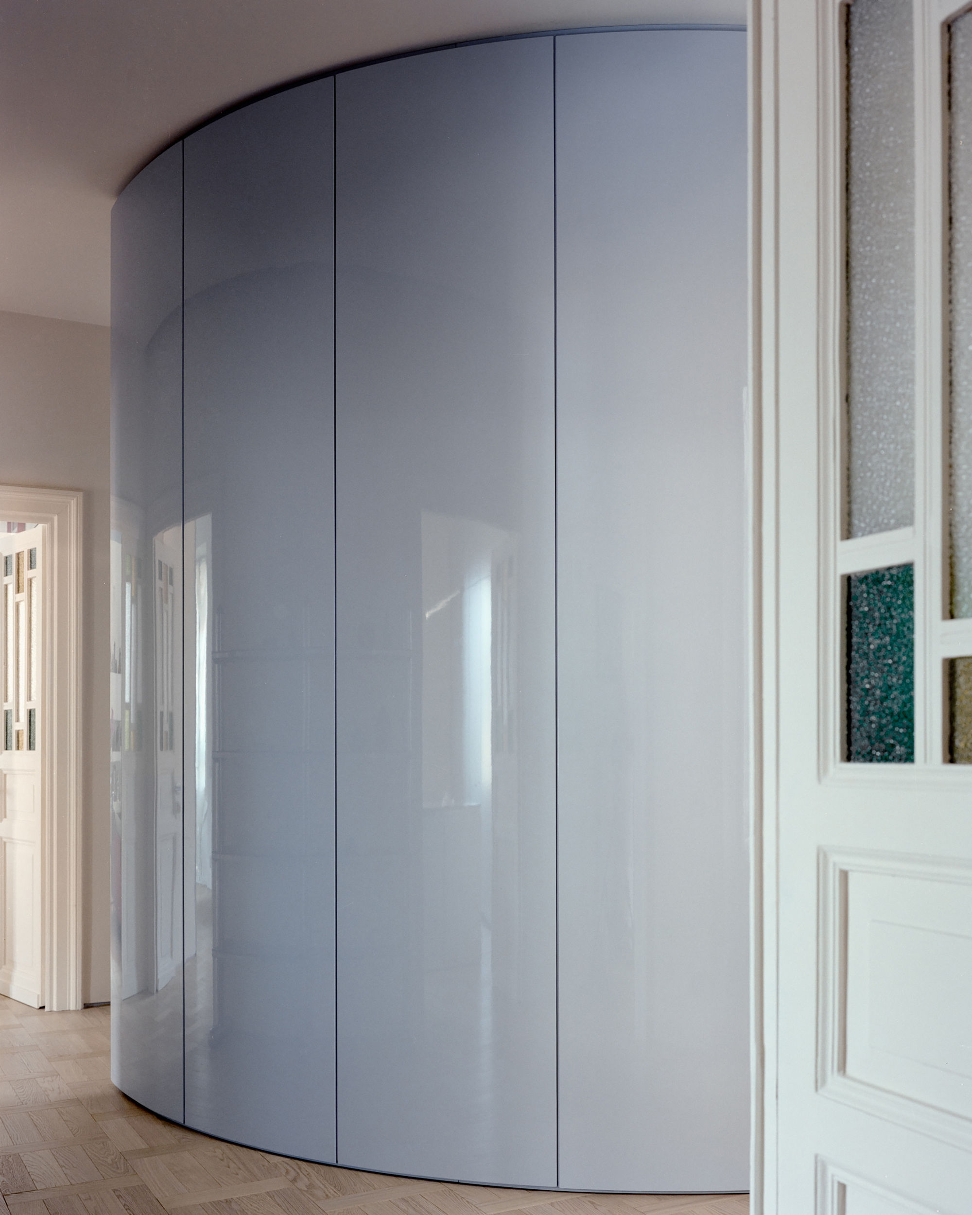 Curved glossy blue-grey cabinet volume in a hallway, next to a white paneled door with textured glass panes