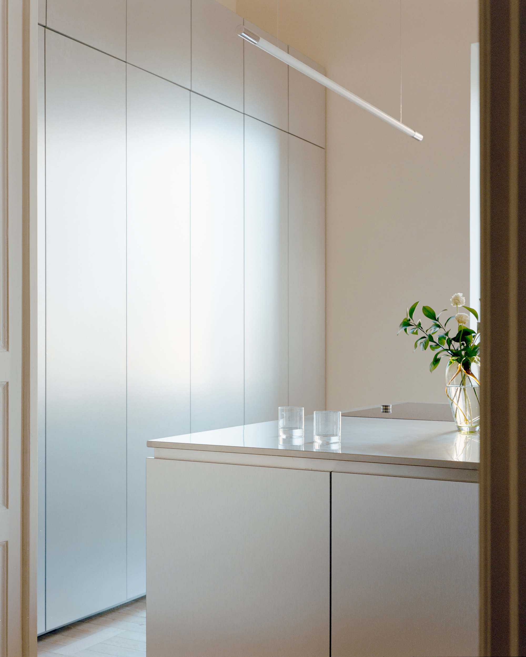 Corner of a steel kitchen island with two glasses and a flower vase, beside frosted tall cabinets under a slim pendant light