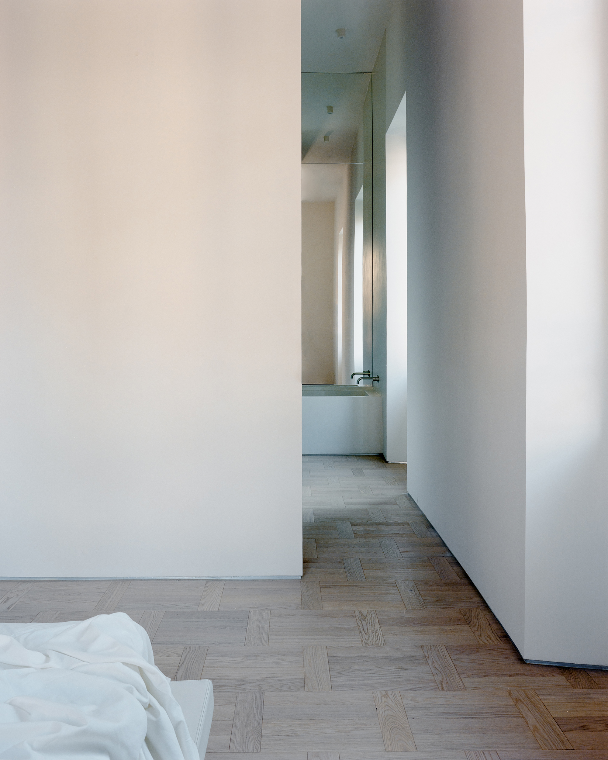 Parquet floor leading past a white wall toward a mirrored bathroom with a basin and wall-mounted taps in soft light