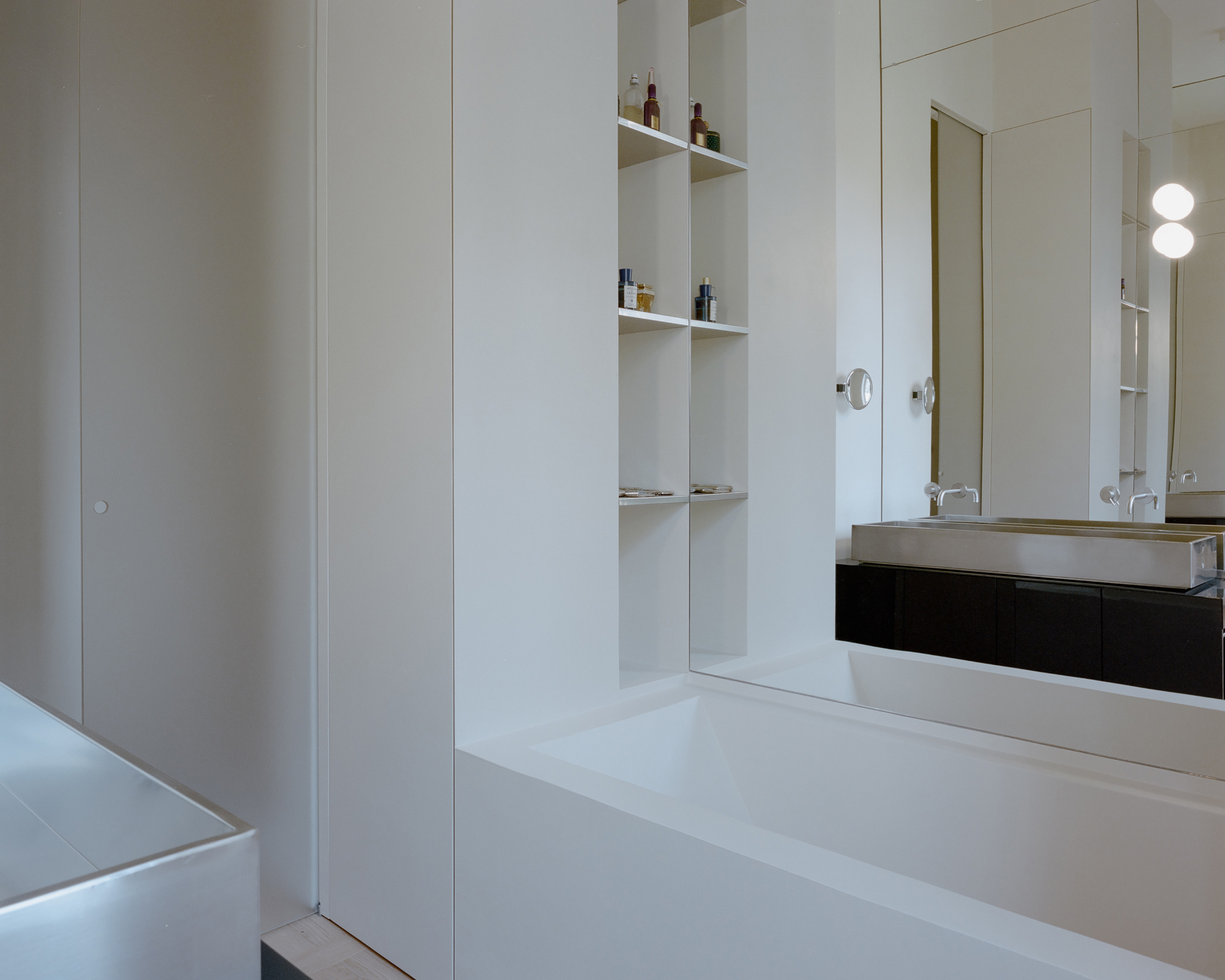 White built-in shelving beside a solid white bathtub, with a stainless steel basin reflected in a large mirror under soft daylight