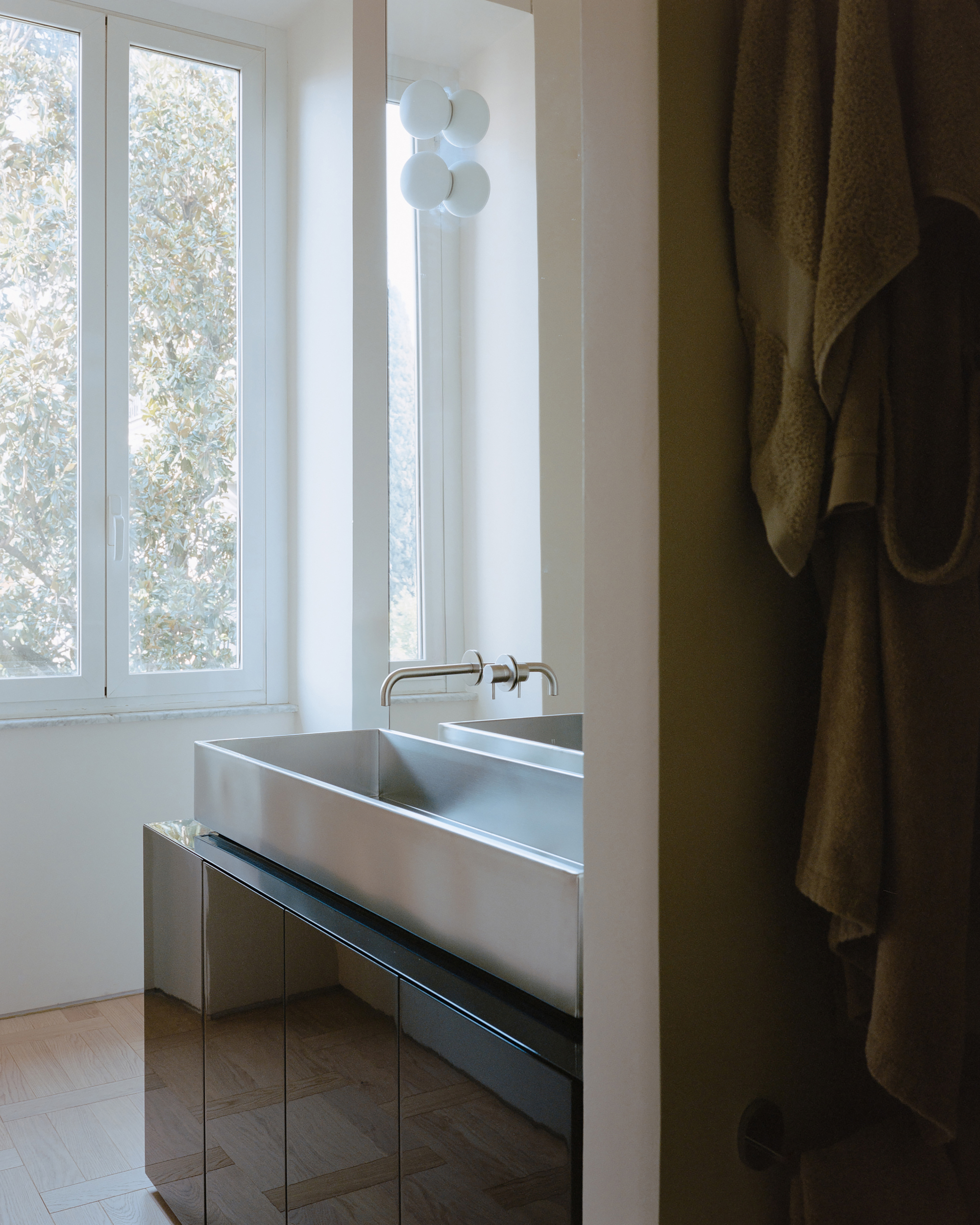 Stainless steel trough sink on a black mirrored vanity beside a tall window filled with sunlit leaves and a hanging robe