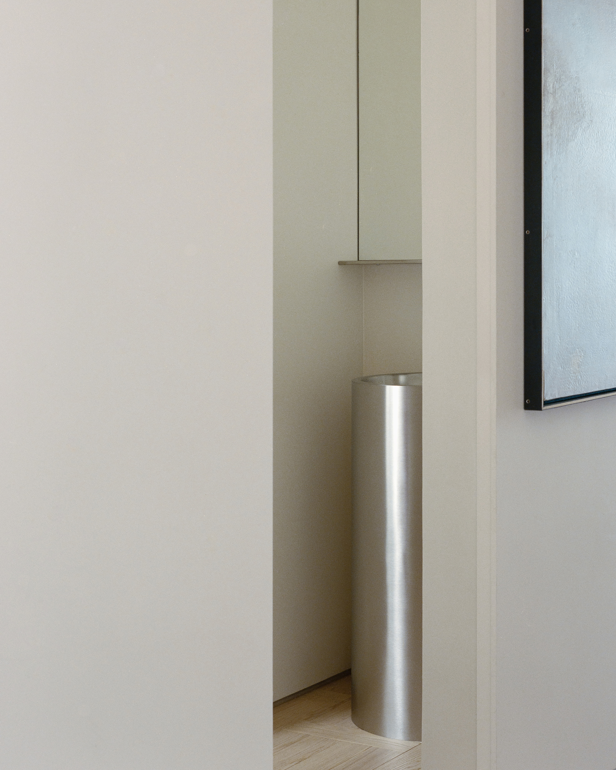 Narrow gap between white walls revealing a cylindrical stainless steel pedestal basin, with a framed painting at right in soft light
