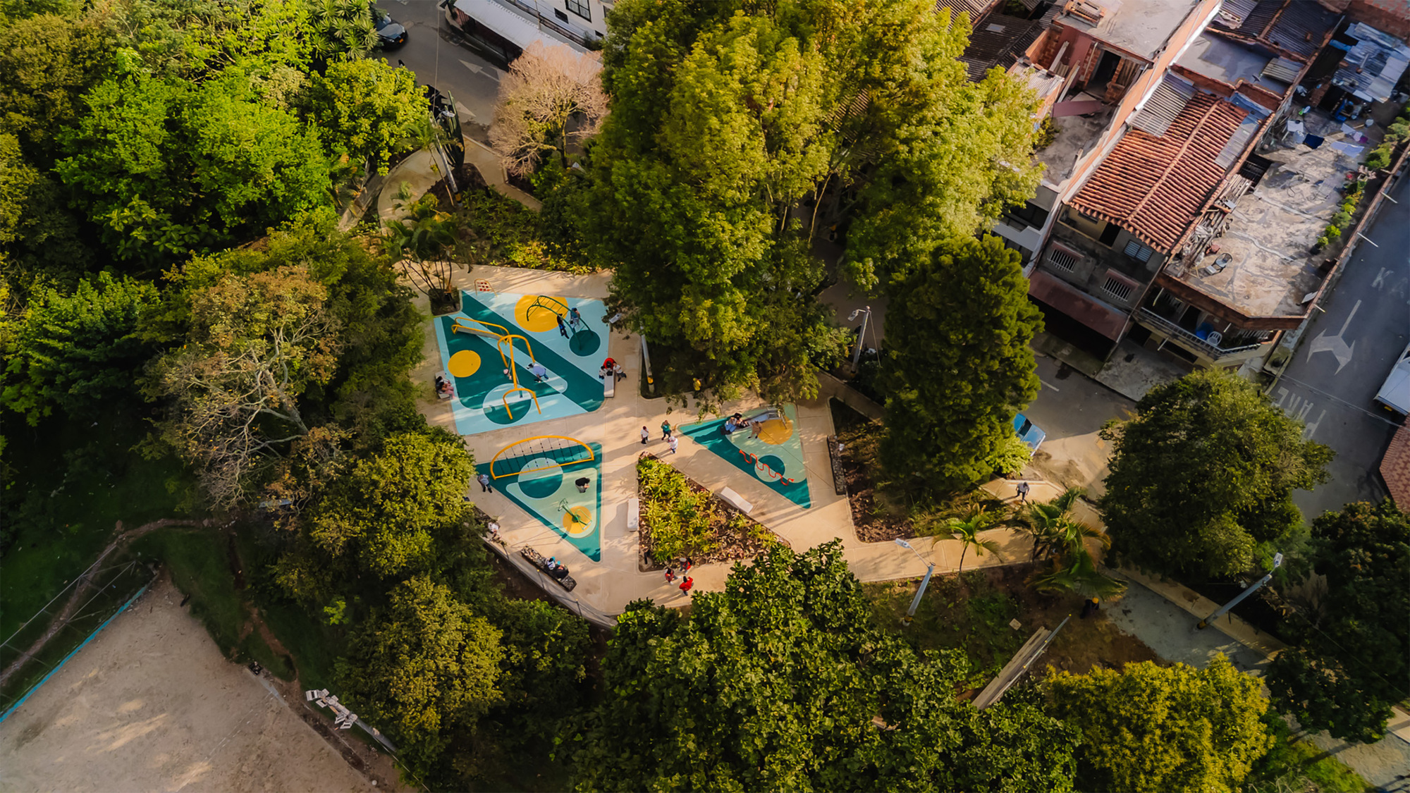 Aerial view of teal and yellow playground surfaces on a paved path, ringed by tree canopy beside red-tiled rooftops