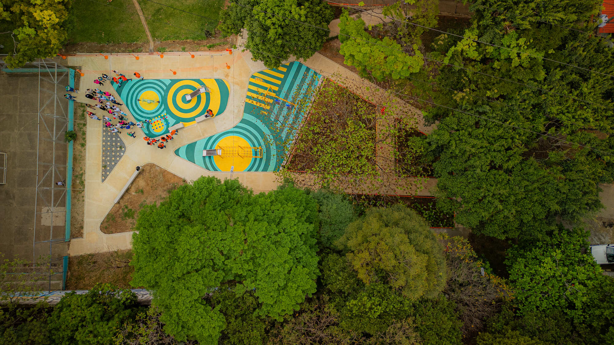 Top-down view of striped teal and yellow play surfaces with a group of children gathered, surrounded by dense green trees
