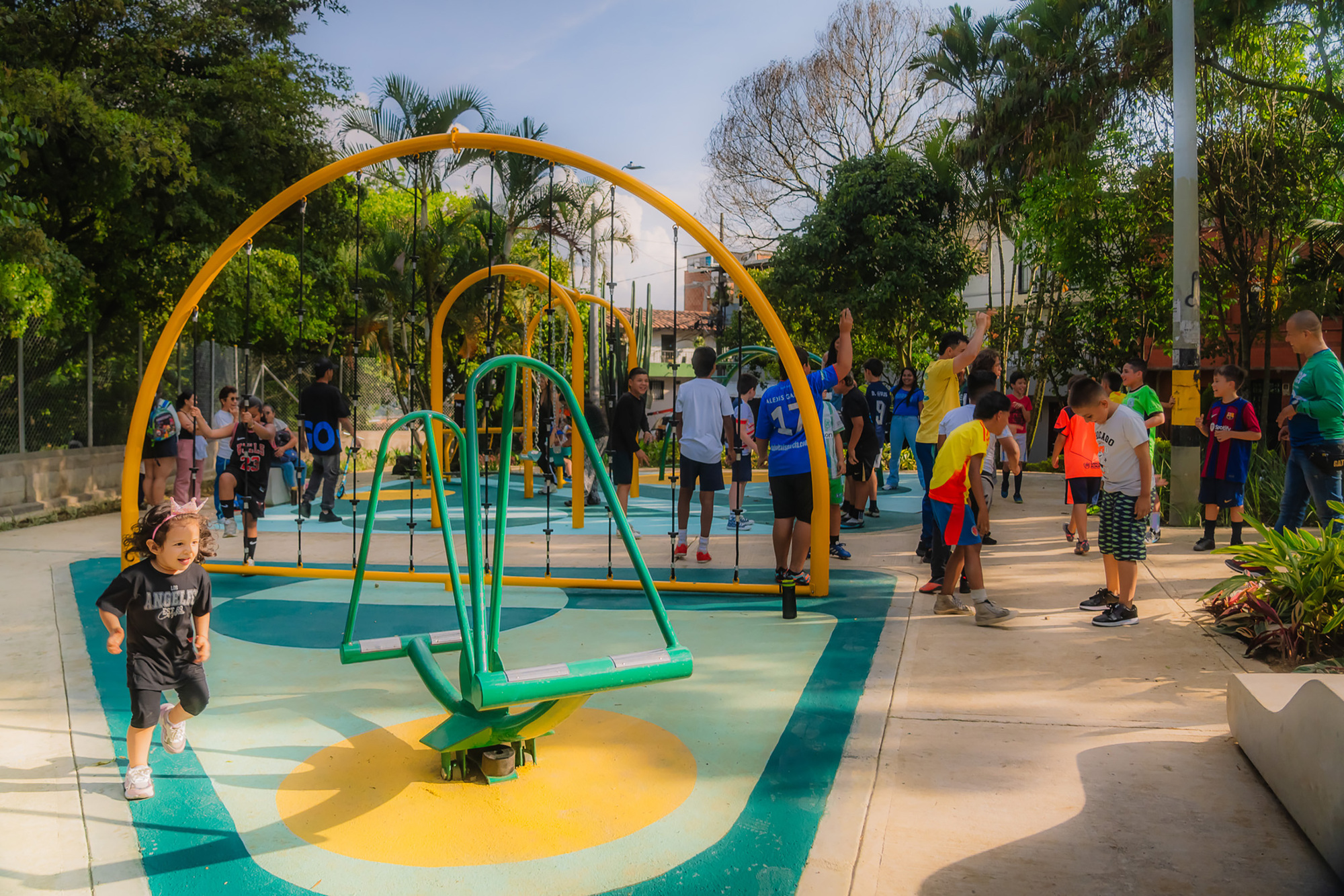 Children running and gathering around a green spinning seat and yellow arches on the teal and yellow play surface in late light