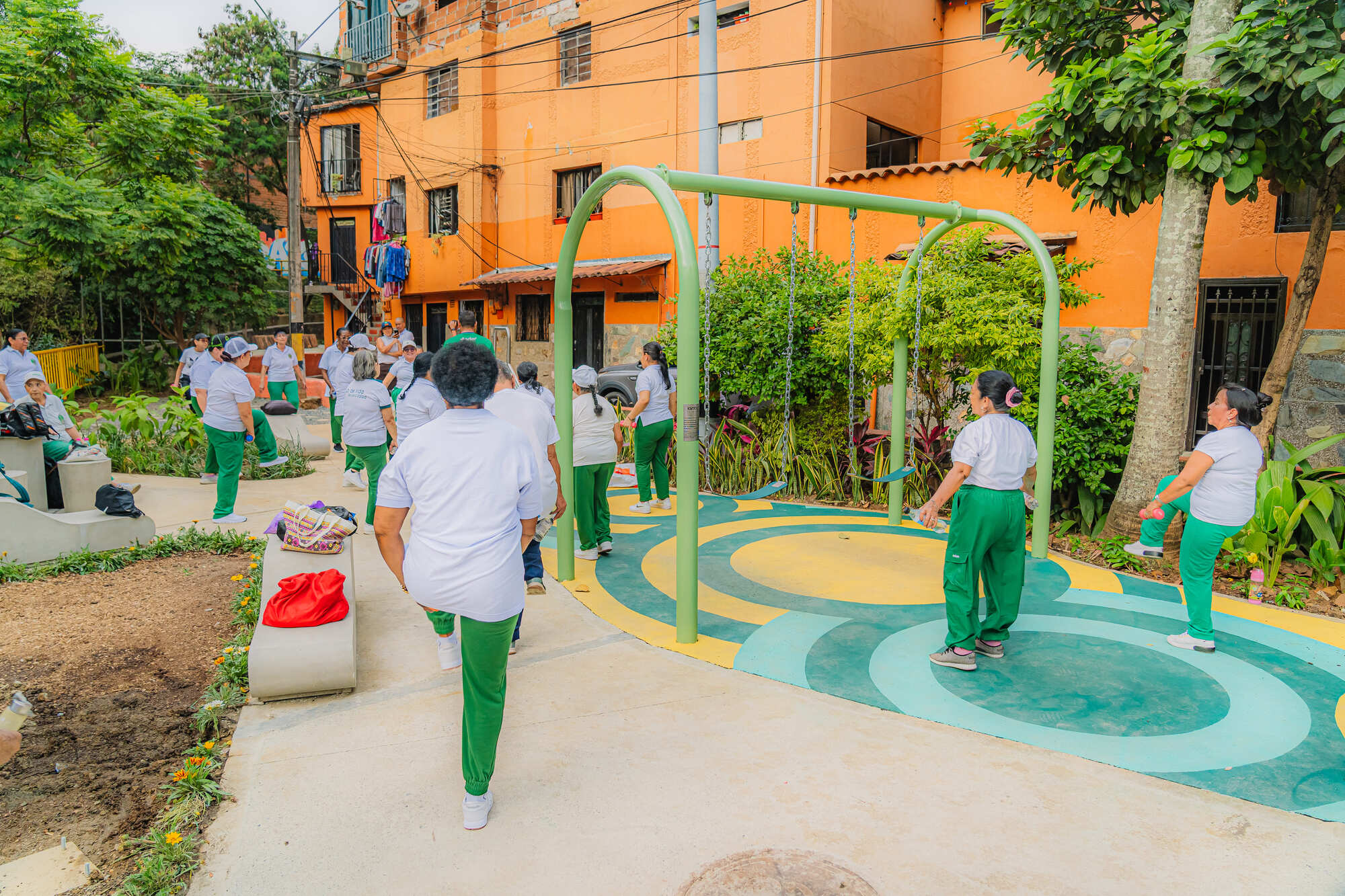 Group in white shirts and green trousers exercising beside green swing frames, with an orange housing facade behind the trees