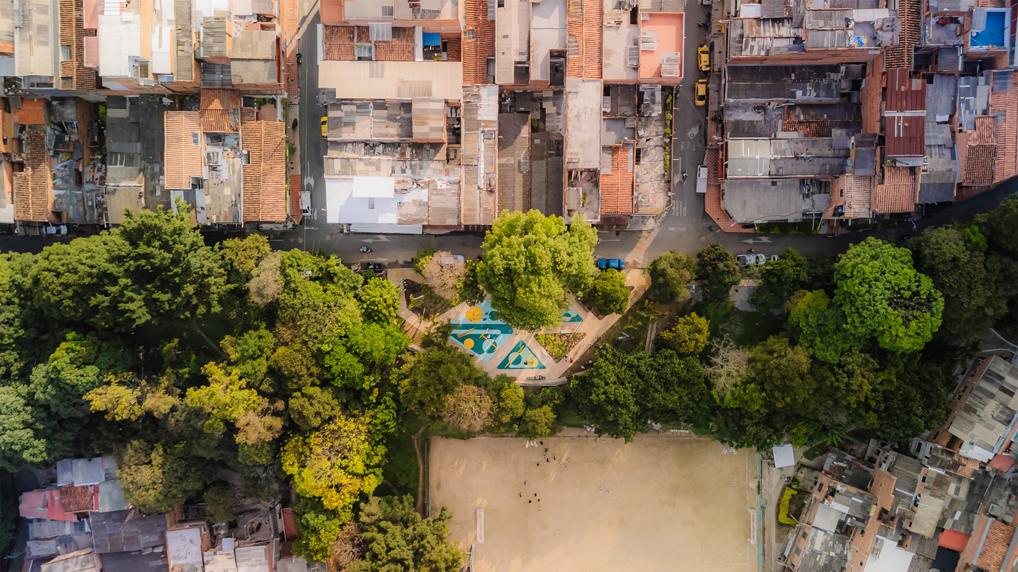 Aerial view of the small playground tucked between dense tree cover, a dirt field and packed rooftops along a street