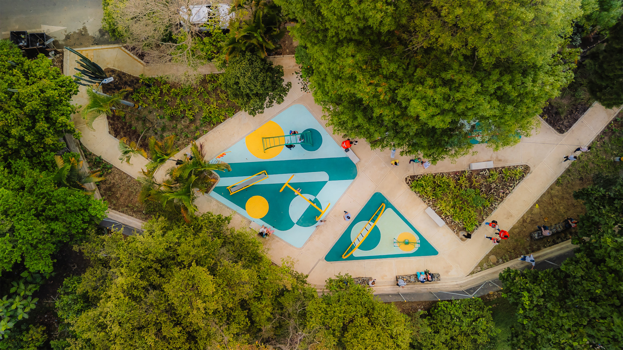 Top-down view of teal and yellow rubber play surfaces with climbing frames and planted triangular beds beneath dense tree canopy
