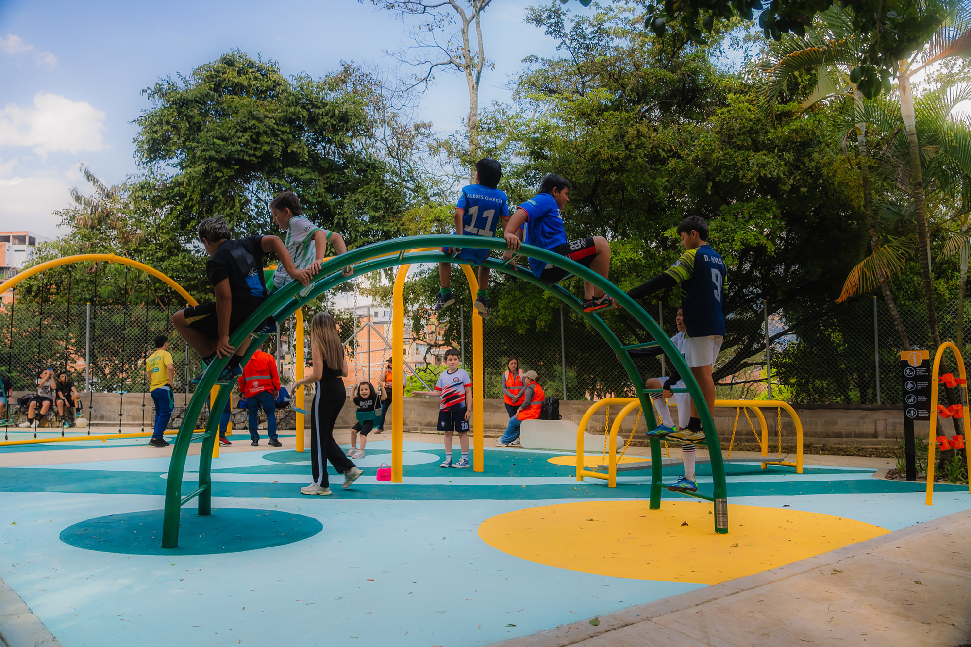 Children climbing a green arched steel frame above a painted teal and yellow play surface under a bright sky