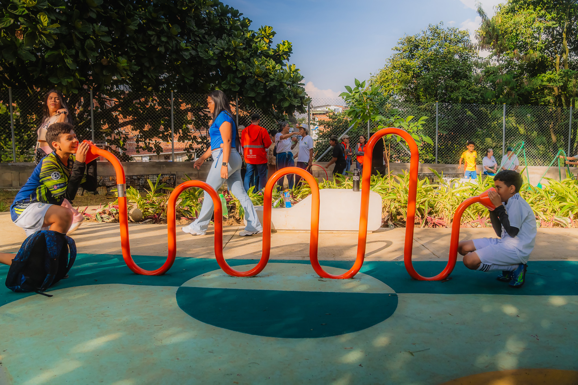 Two boys speaking into orange serpentine tube telephones while adults walk past in dappled afternoon sunlight