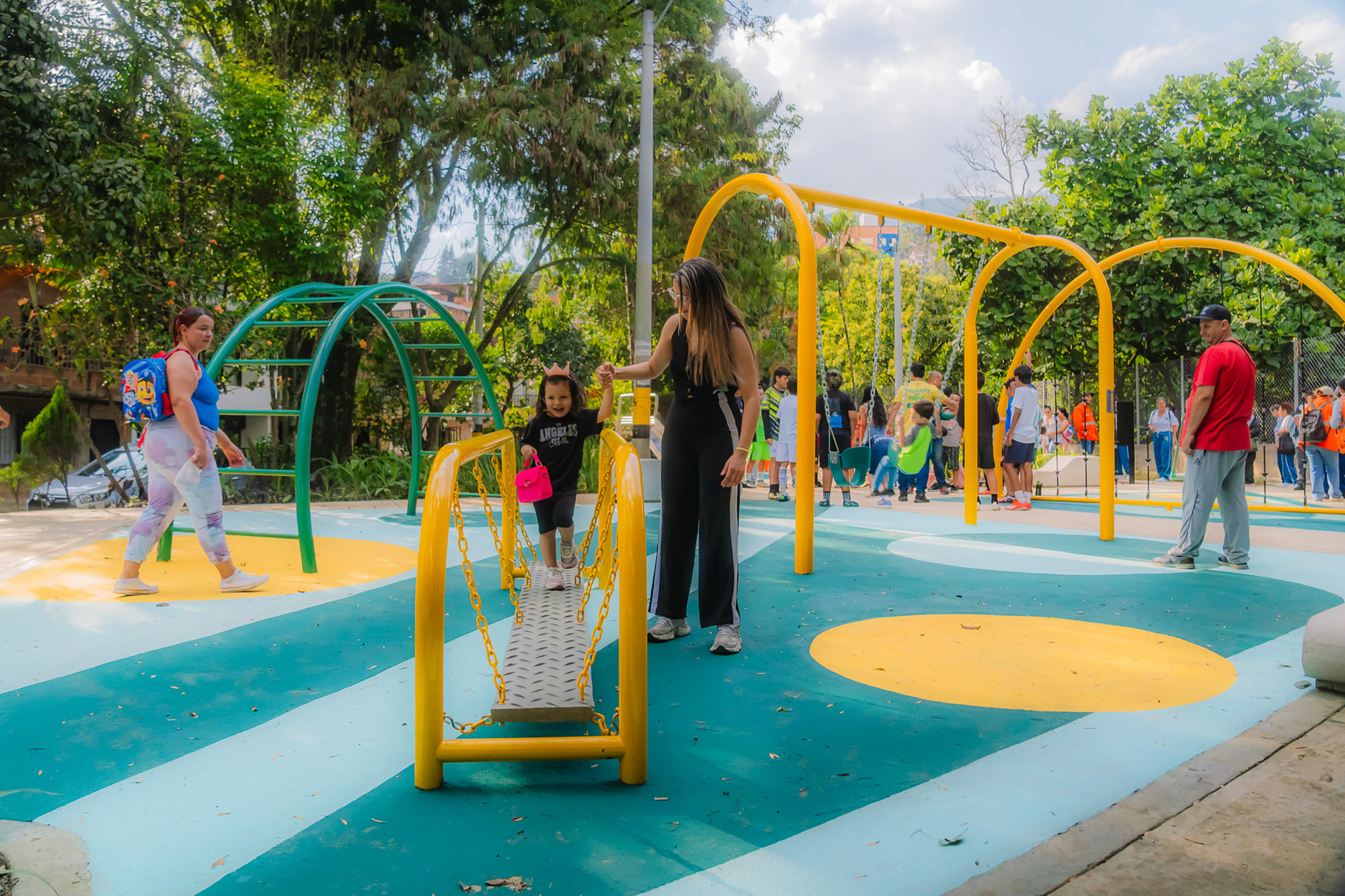 Yellow chain bridge and arched swing frames on a teal play surface with a child crossing and a woman holding her hand