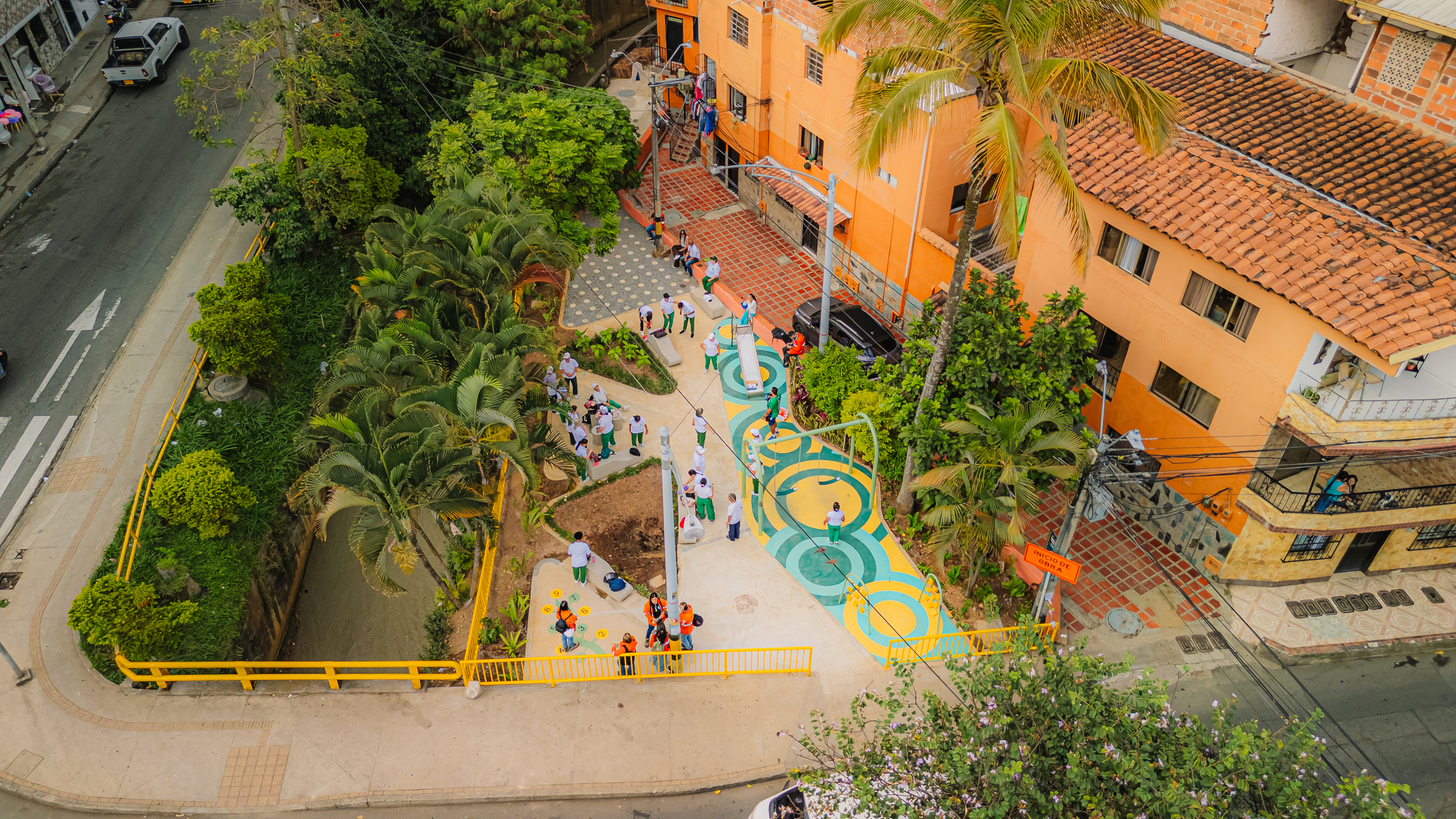 Oblique aerial view of a small play plaza with painted concentric circles, palms and a yellow railing beside orange houses