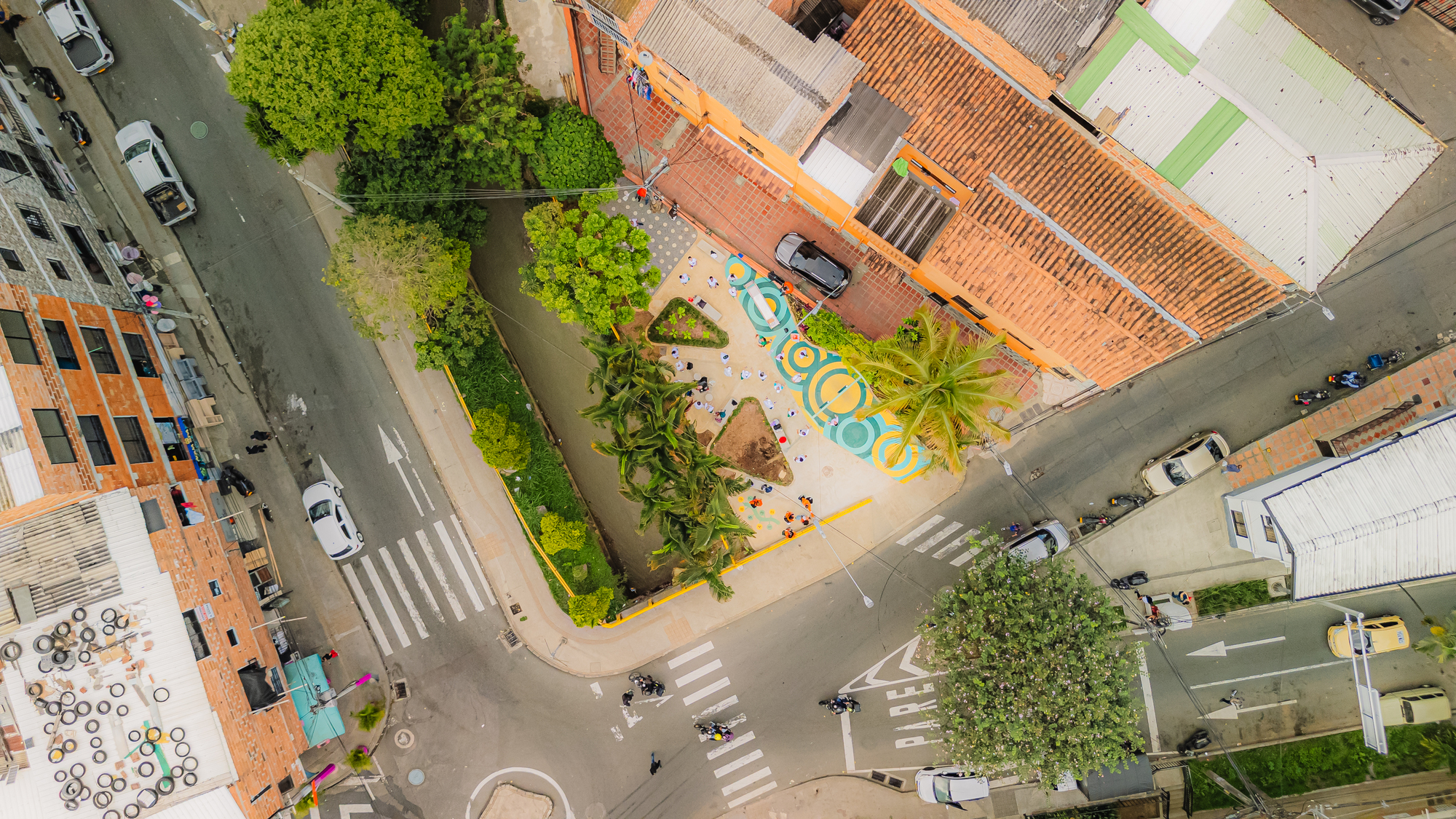 Aerial view of a narrow corner plaza with painted circles and palms between clay-tiled roofs and a street junction with crosswalks