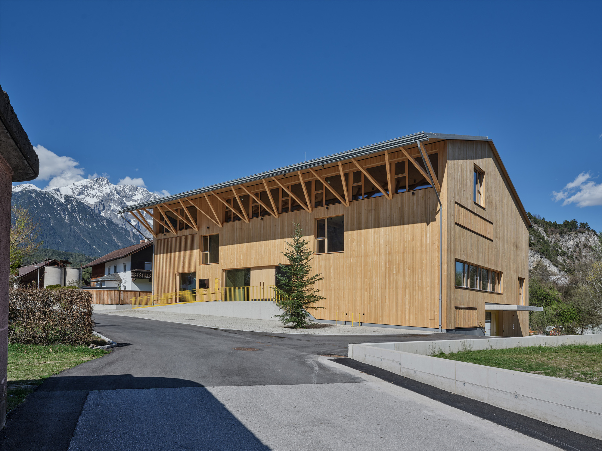 Timber-clad building with exposed diagonal roof brackets and a lone spruce, set against snow-capped peaks under a clear blue sky