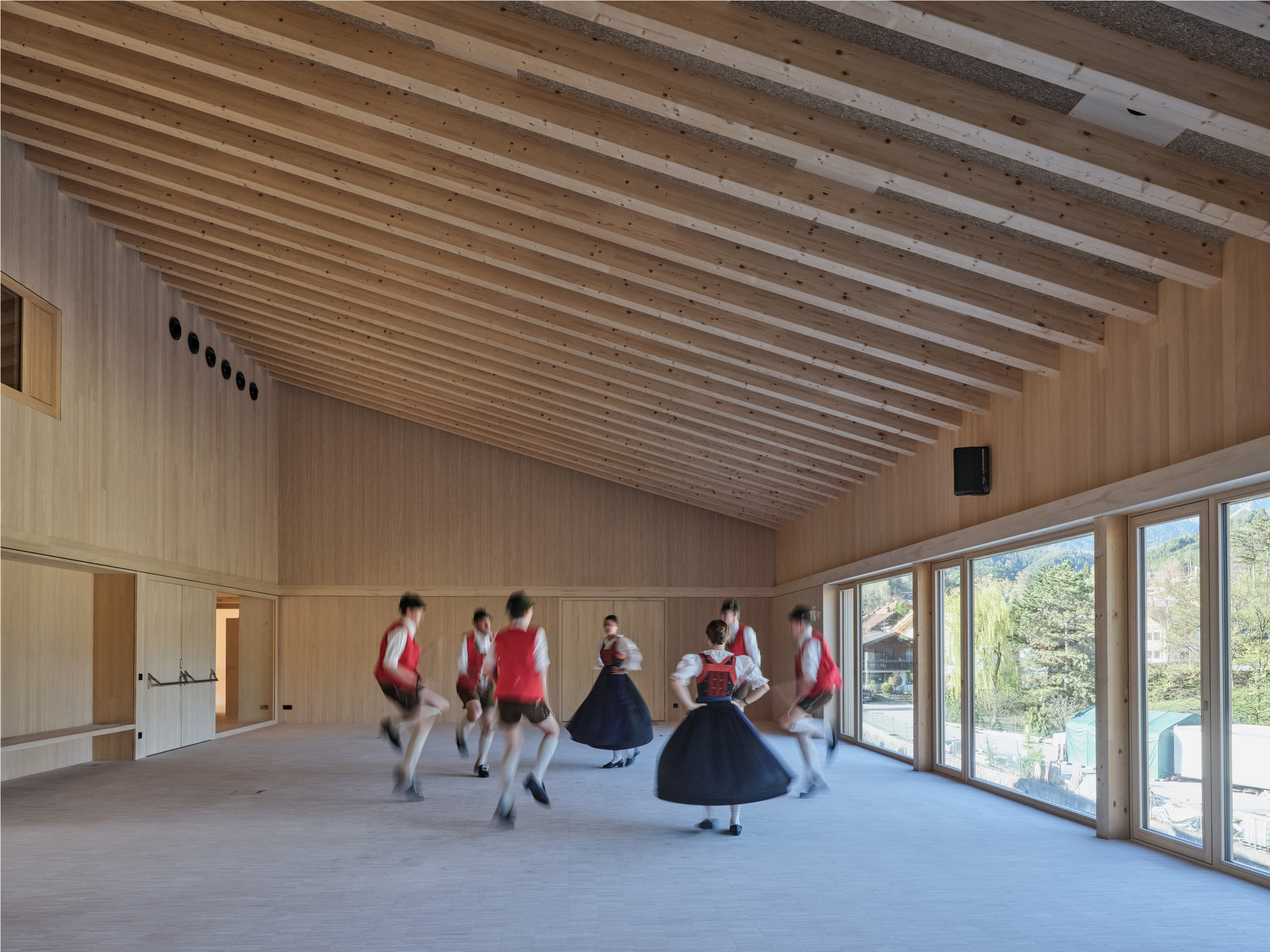 Dancers in traditional red vests and dark skirts whirling across a timber hall beneath a sloped ceiling of exposed rafters