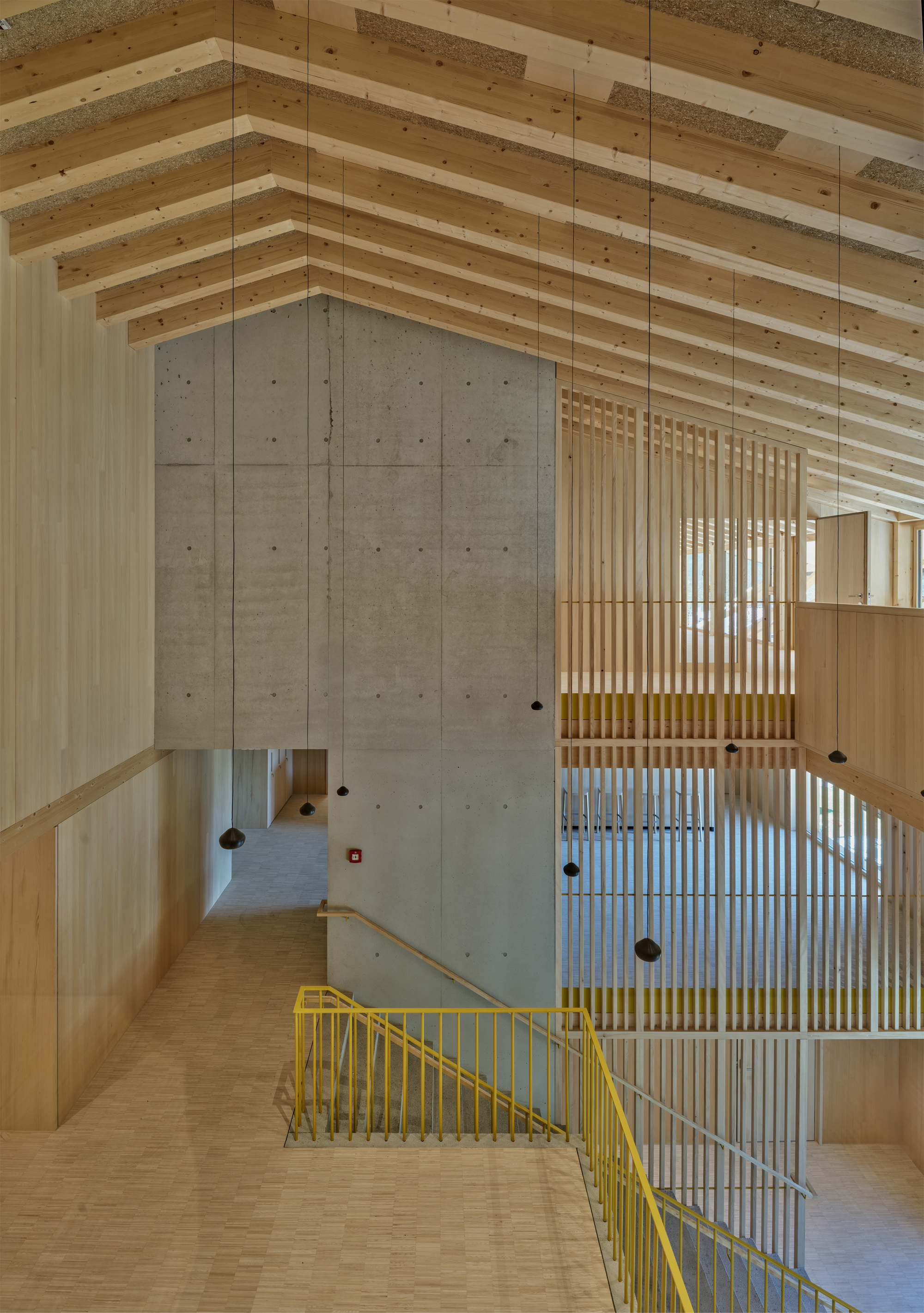 View down a stairwell with yellow railings, a board-marked concrete wall and vertical timber slats beneath a pitched beam ceiling