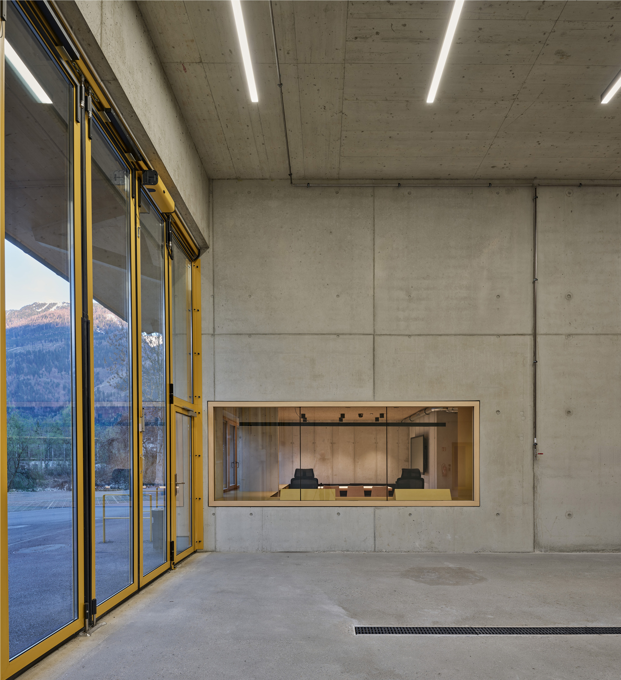 Concrete room with a yellow-framed glass wall and a small timber-lined window onto a meeting room, with mountains reflected in the glass