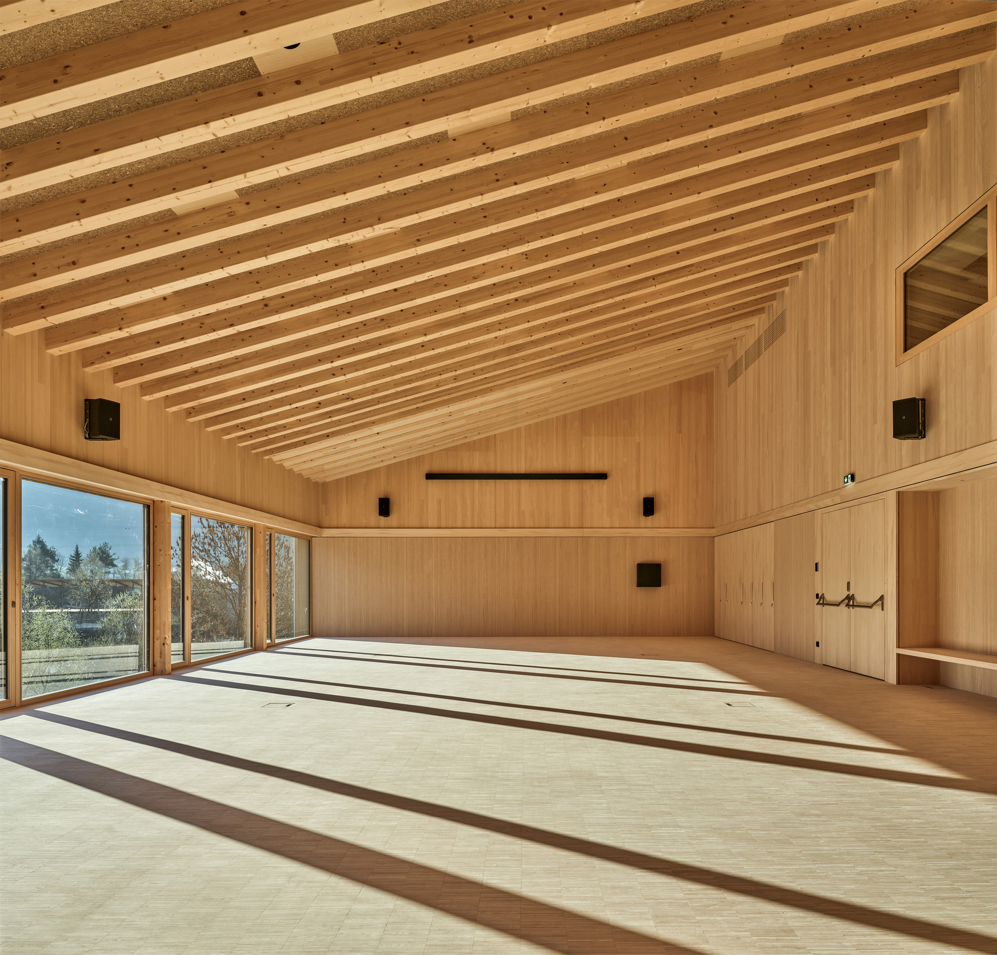 Empty timber hall with a pitched rafter ceiling and long bands of sunlight and shadow falling across the floor