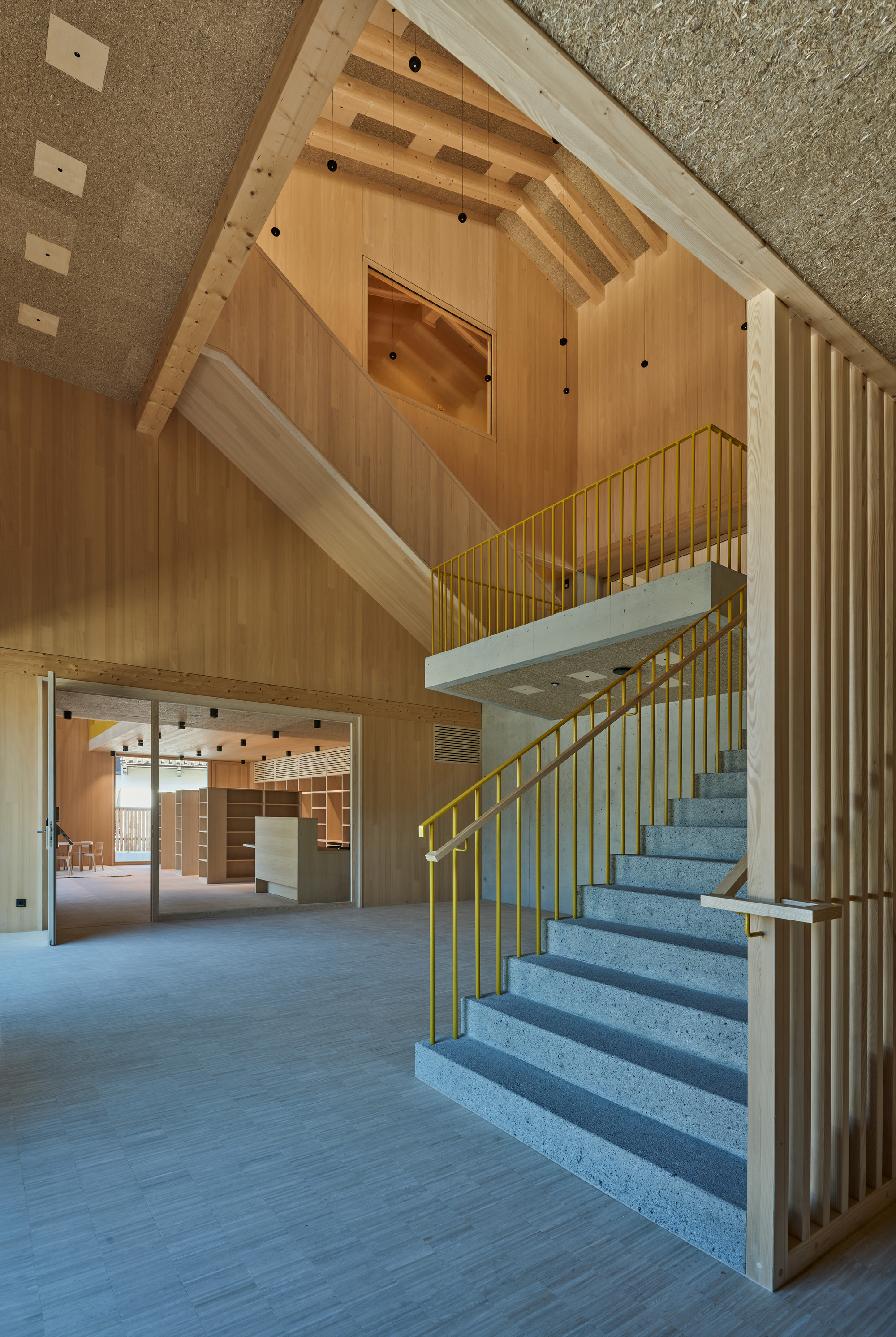Stone-stepped staircase with yellow railings rising through a tall timber-lined hall, with a slatted screen at right and a library beyond