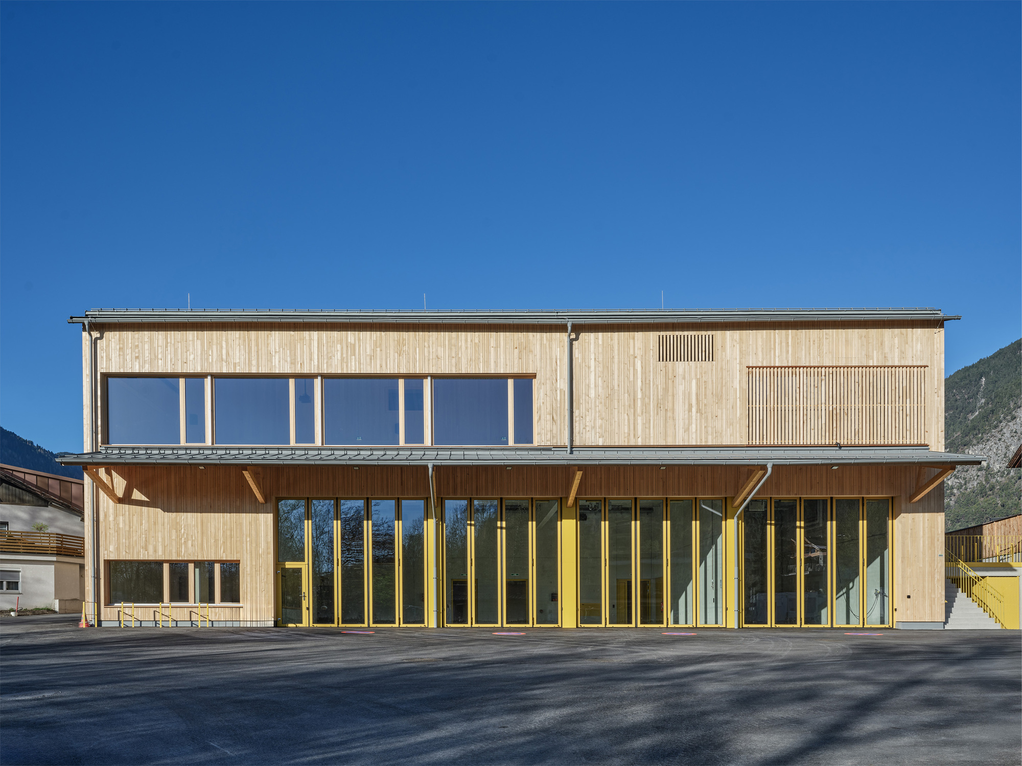 Timber-clad front facade with yellow-framed folding glass doors beneath a deep canopy, set against a clear blue sky and mountain slope