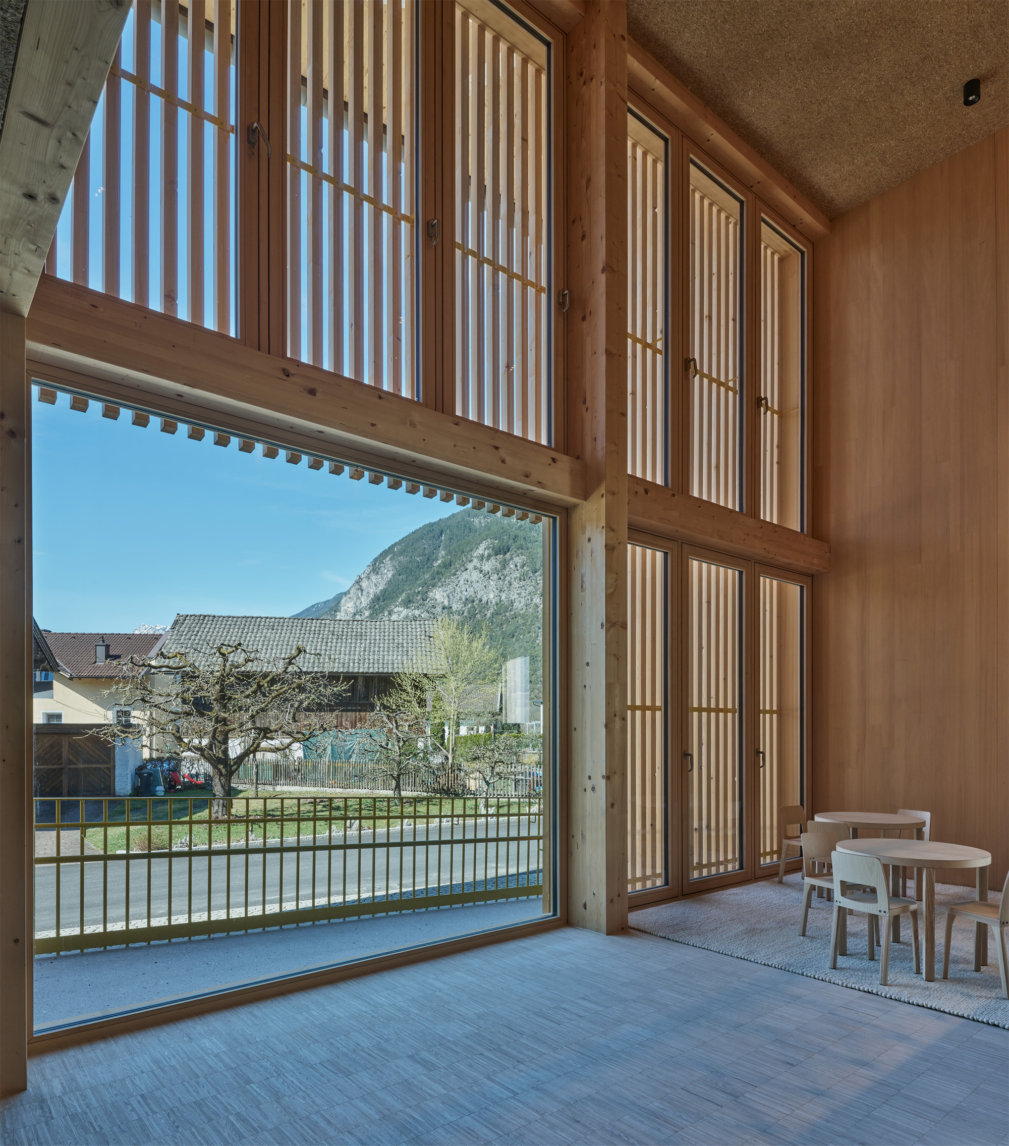 Double-height timber room with tall slatted windows and a large glazed opening framing a mountain, with children's tables at right